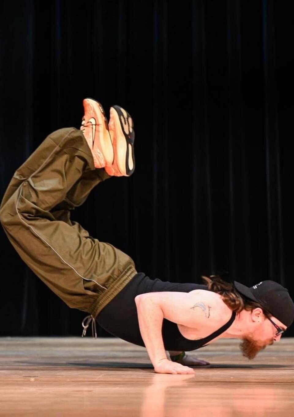 Man wearing a black tank top, black cap, and glasses performing a yoga pose called 'Forearm Balance' on a wooden floor with a black curtain in the background, as part of a hip hop dance performance.