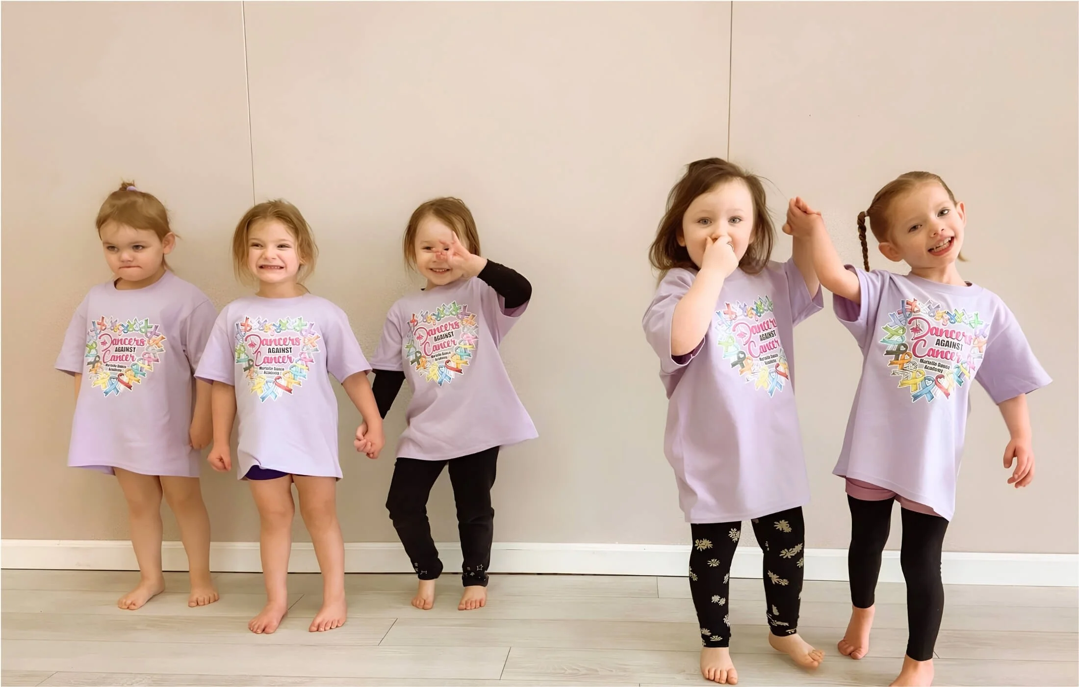 Five young girls standing barefoot against a beige wall, wearing matching purple T-shirts with a colorful ribbon design and 'Dancers Against Cancer' written on the front.