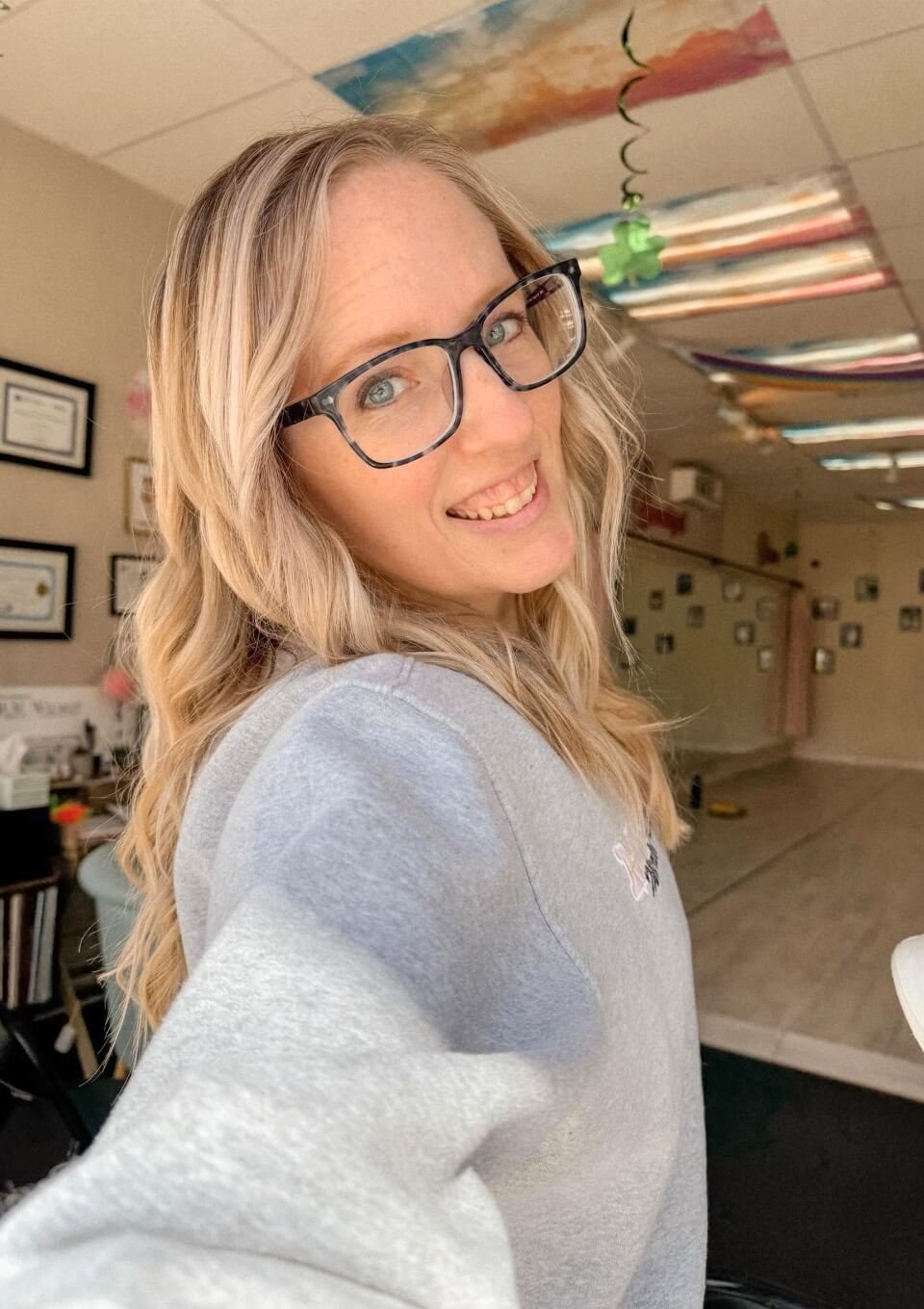 Miss Kate, owner of Martelle Dance Academy with blonde hair wearing glasses and a gray sweatshirt, smiling and taking a selfie inside her dance studio.