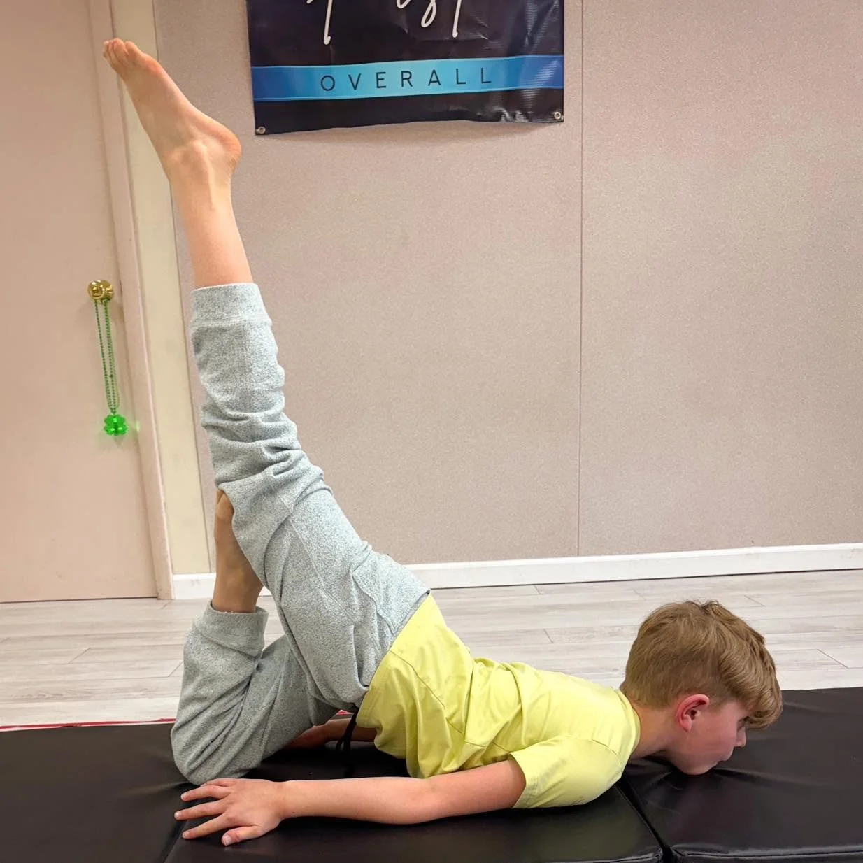 A young boy with red hair in a yellow shirt and gray sweatpants is lying on his stomach on a black exercise mat. He has one leg bent and raised with his foot pointing towards the ceiling, while the other leg is bent underneath him.