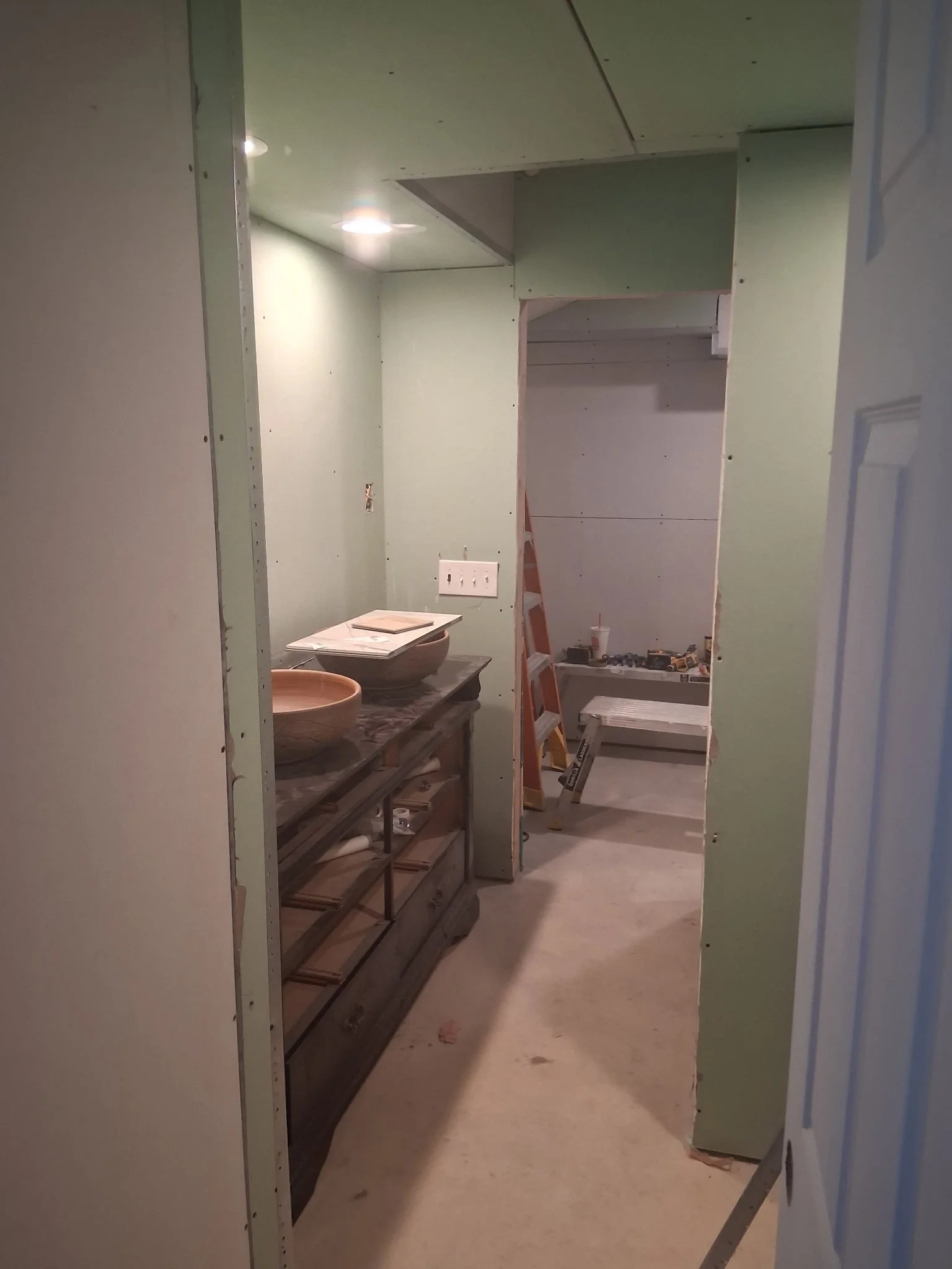 Bathroom under construction with green drywall, a wooden vanity with two vessel sinks, a ladder, and construction tools in the background.