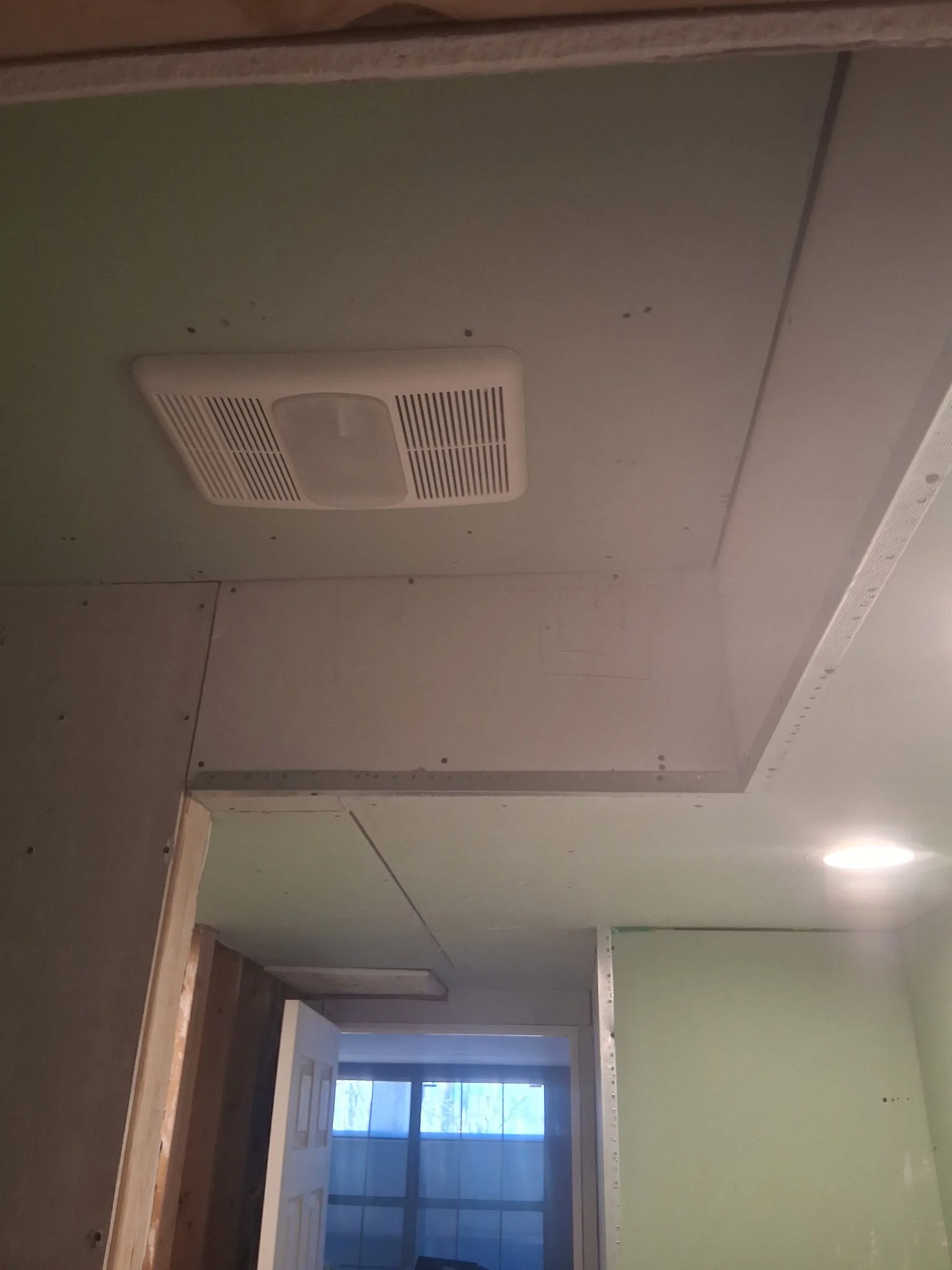 Construction site interior with unfinished drywall, ceiling vent, and a partially open door leading outside with a window showing trees.