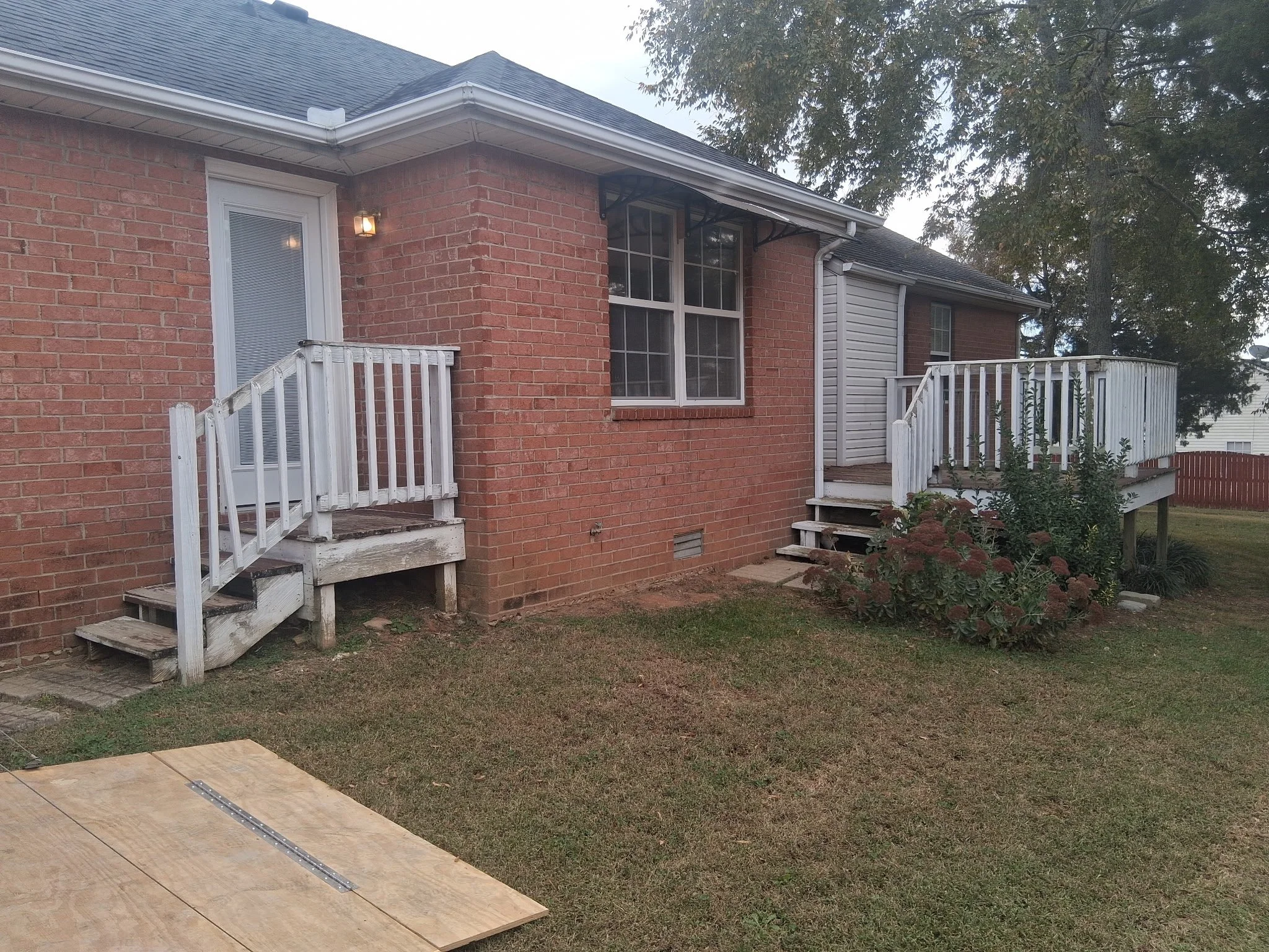 Back of a brick house with two small decks, one on each side, leading to doors. The decks have white railings and steps, and the house has a large window between them. There is a small light fixture on the wall near the left deck. The yard has grass, some plants, and a plywood sheet on the ground.