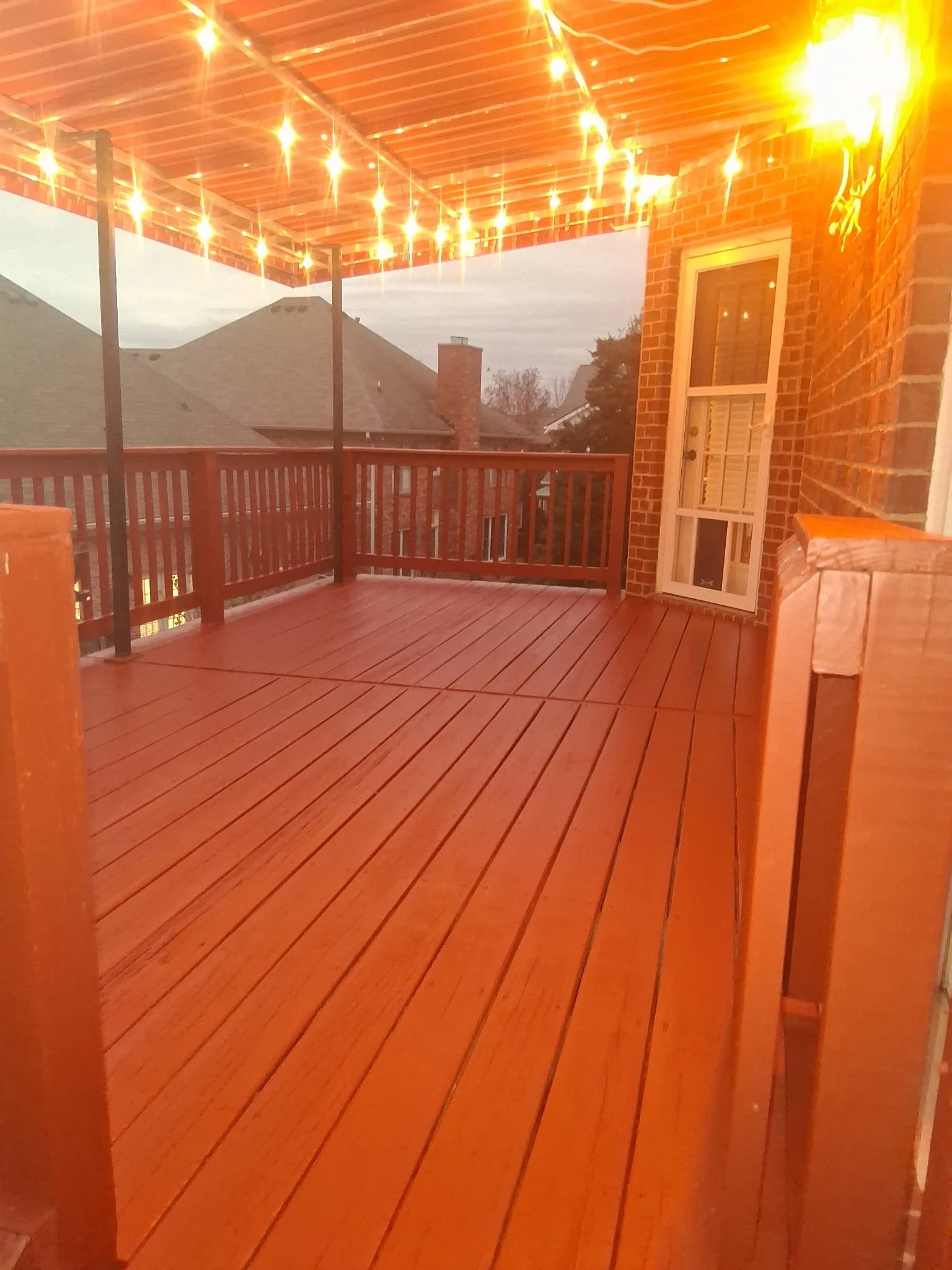Wooden porch or deck with string lights hanging overhead, brick house wall, door with glass window, and neighboring rooftops visible in the background during dusk or evening.