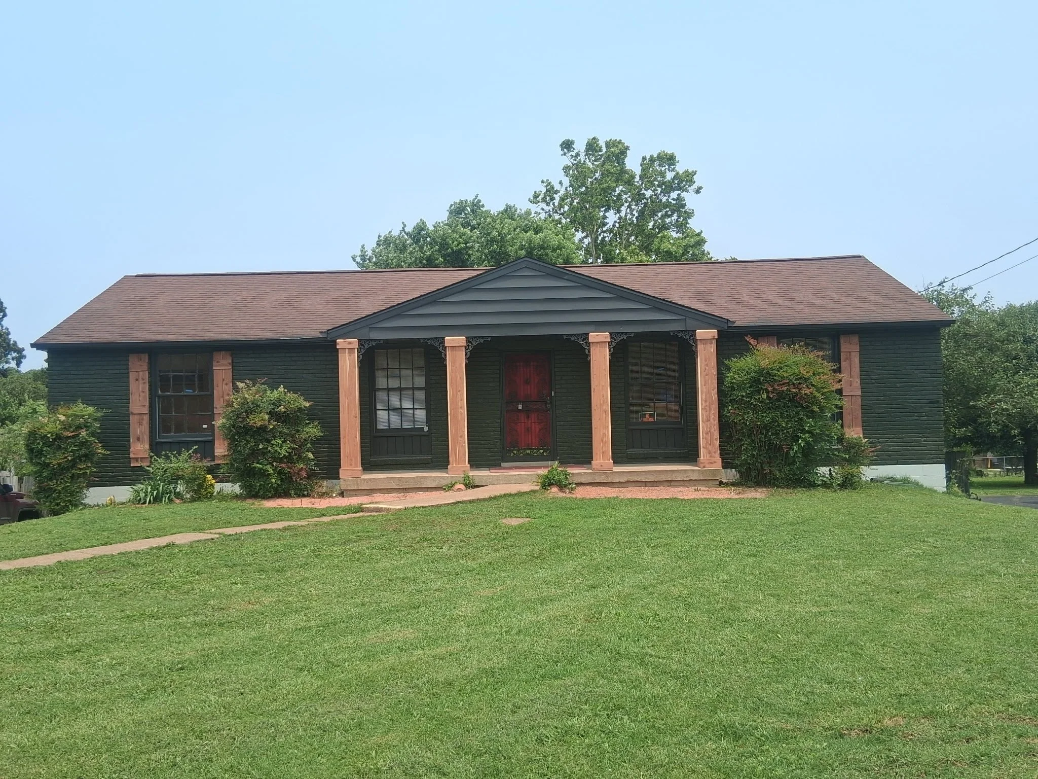 A dark green single-story house with a brown shingle roof, four pinkish wooden columns supporting a small portico with decorative brackets, and a red front door. There are windows with black frames on either side of the door, and shrubs with green foliage and some flowers in front of the house. The house is on a raised lawn with a walkway leading to the front steps, and there are trees in the background.