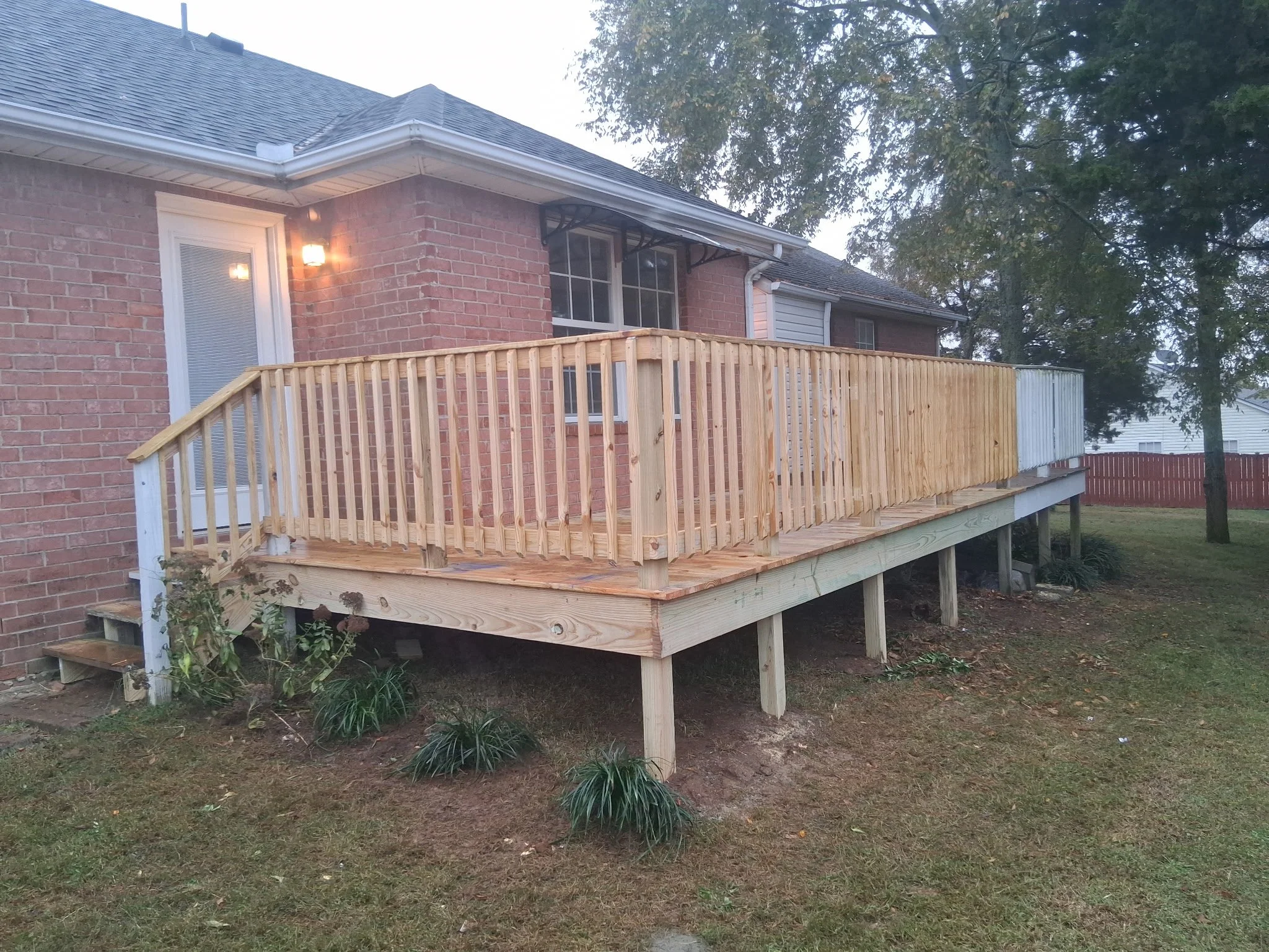 A newly built wooden deck attached to a brick house with a white door and window. The deck has a railing and is elevated with support posts underneath. There are small plants and grass in the yard, with trees visible in the background.