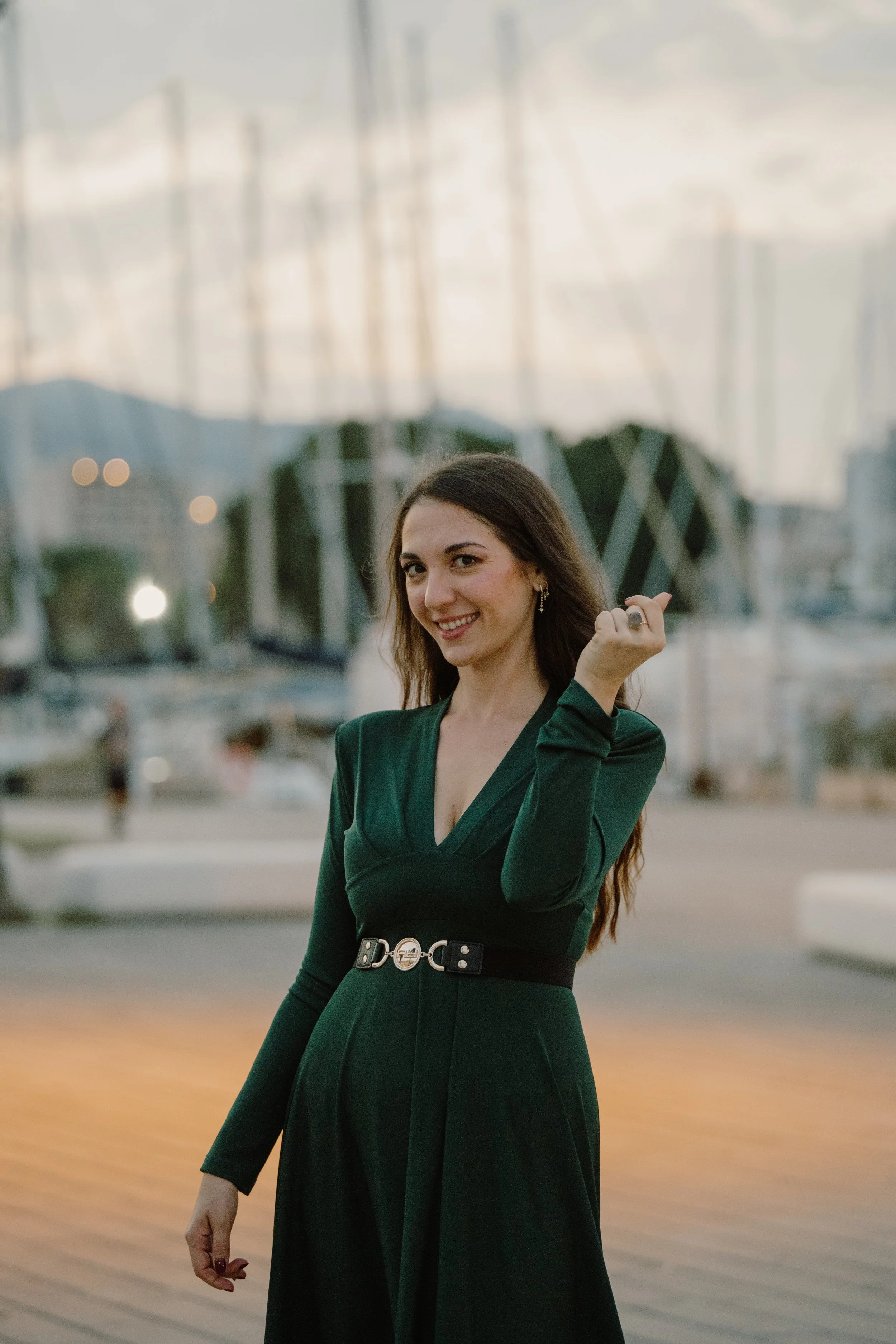Donna sorridente con capelli morbidi, indossa un vestito verde scuro con cintura e mostra il pollice verso in un porticciolo con barche e luci soffuse sullo sfondo durante il tramonto.