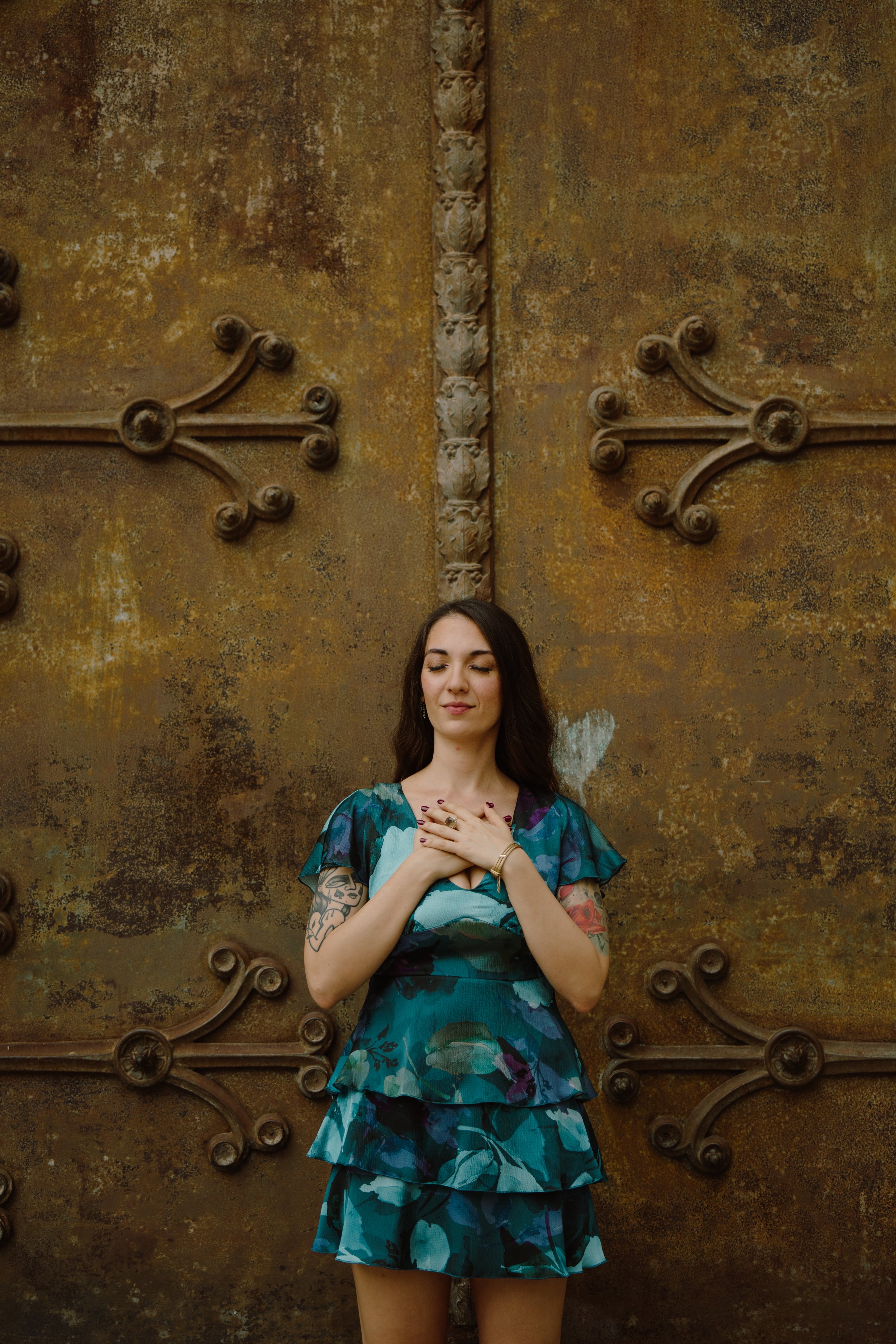 Una donna con capelli lunghi castani, tatuaggi sulle braccia, vestito blu con stampa floreale, si tiene il cuore con le mani e ha gli occhi chiusi davanti a una grande porta di ferro arrugginita.