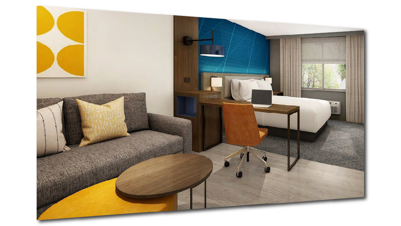 A modern hotel guest room - Comfort Suites, Rise and Shine scheme - with a gray sofa, yellow pillow, abstract yellow artwork, a wooden coffee table, a workspace with a computer and a brown leather chair, a bed with white linens, a blue accent wall.