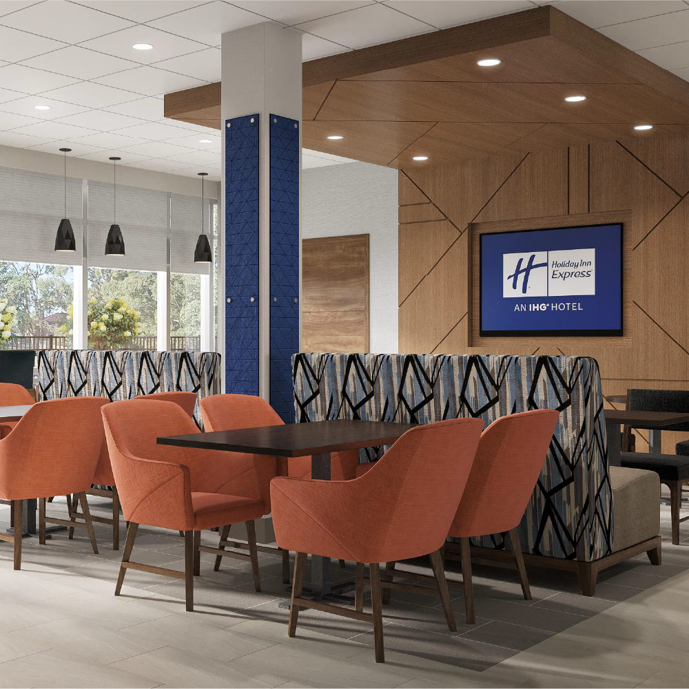 Holiday Inn Express Formula Blue 2.0 SRD hotel lobby area with orange chairs around dark wood tables, patterned booth seating, large windows with blinds, a mounted TV displaying the Holiday Inn Express logo, and modern ceiling and lighting fixtures.