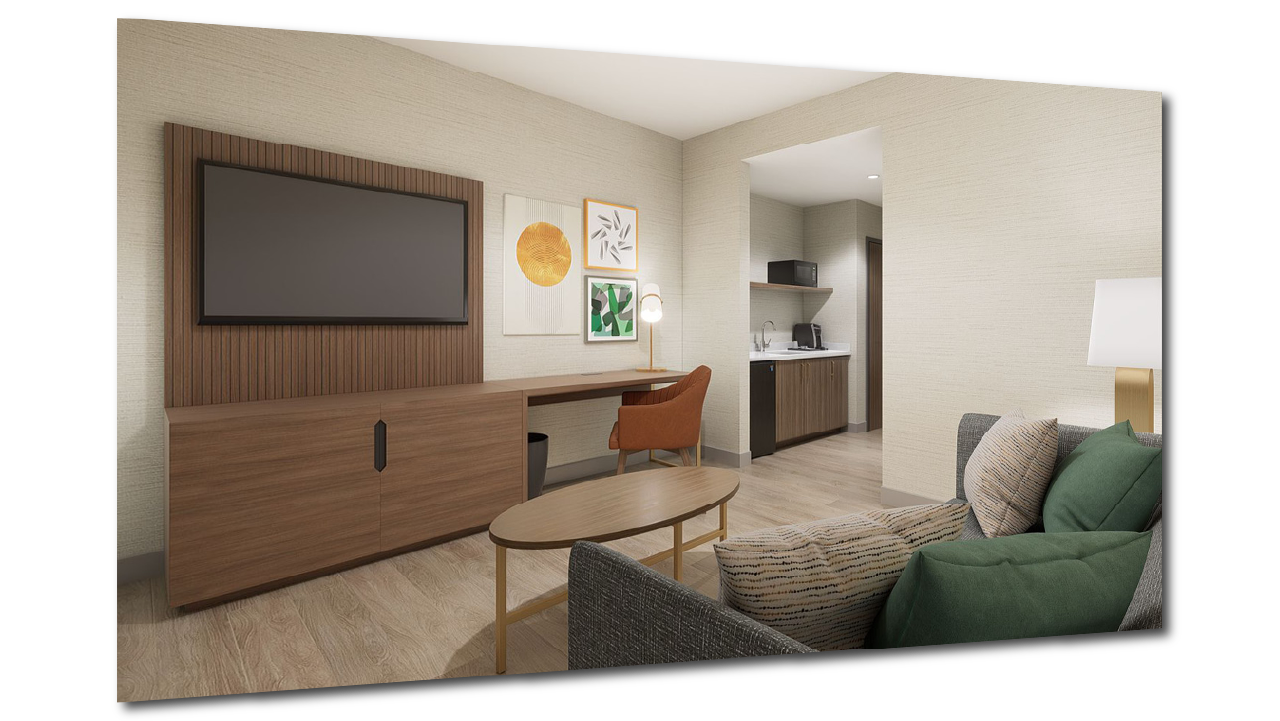 View of a modern Holiday Inn H5 prototype hotel guest room with a wall-mounted flat-screen TV, abstract art on the wall, a desk with an orange chair, a sofa with patterned and green pillows, a wooden coffee table, and a kitchenette in the background.
