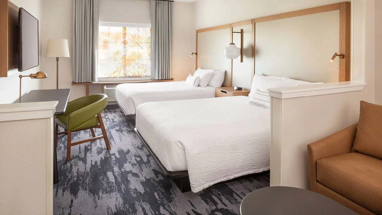 Modern prototypical guest room by Fairfield Inn and Suites by Marriott, with two queen beds, a window with curtains, a TV on the wall, a desk with a green chair, a brown sofa, and a lamp.