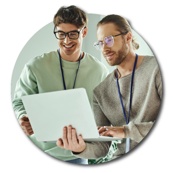 Two designers looking at a laptop together in an office setting, smiling, one wearing glasses and a gray sweater, the other in a light green shirt. This is a scene from a medium-sized interior design studio.