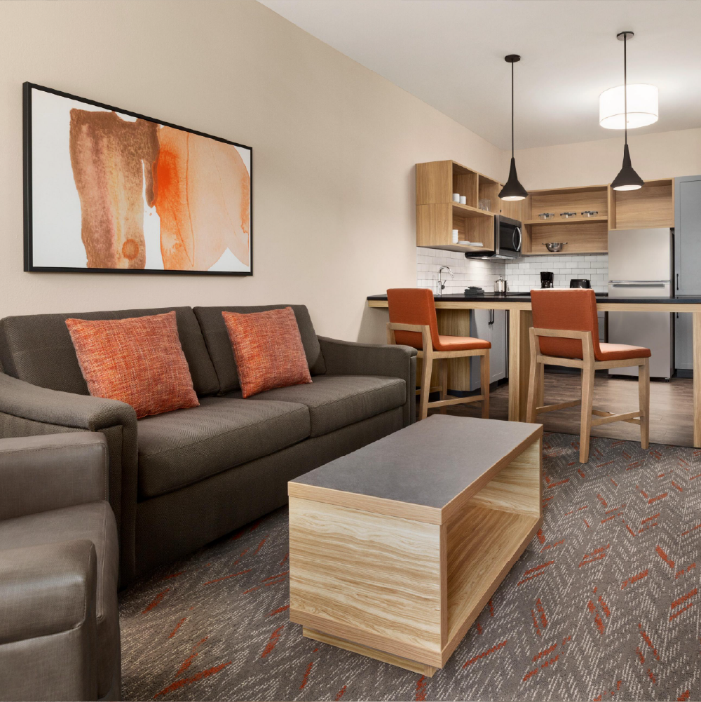 Living room and kitchenette in a modern prototypical hotel - Candlewood Suites Beacon scheme. Gray sofa, orange pillows, abstract wall art, wooden coffee table, two orange bar stools, open shelving, white subway tile backsplash.