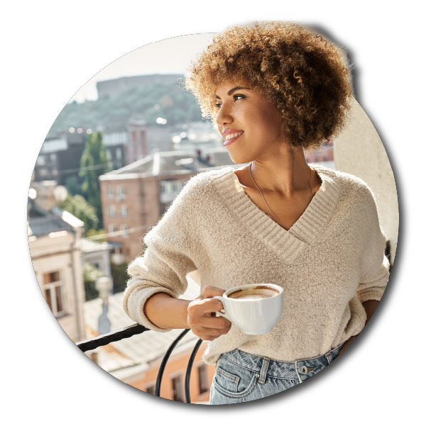 A happy guest, this is a young woman with curly hair holding a coffee cup on a hotel balcony with city buildings in the background.