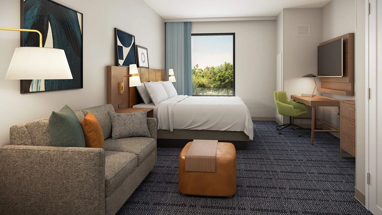A Staybridge by IHG modern hotel room with a bed, a beige sofa with pillows, artwork on the walls, a window with view of trees, a wooden desk with an attached TV, and a green chair.