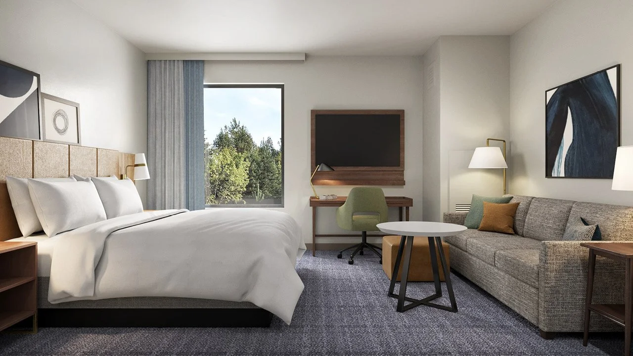 Staybridge Suites by IHG prototypical hotel room with a king-sized bed, a large window with curtains, a flat-screen TV mounted on the wall, a desk with a green chair, a sofa with cushions, a round coffee table, and modern lamps.