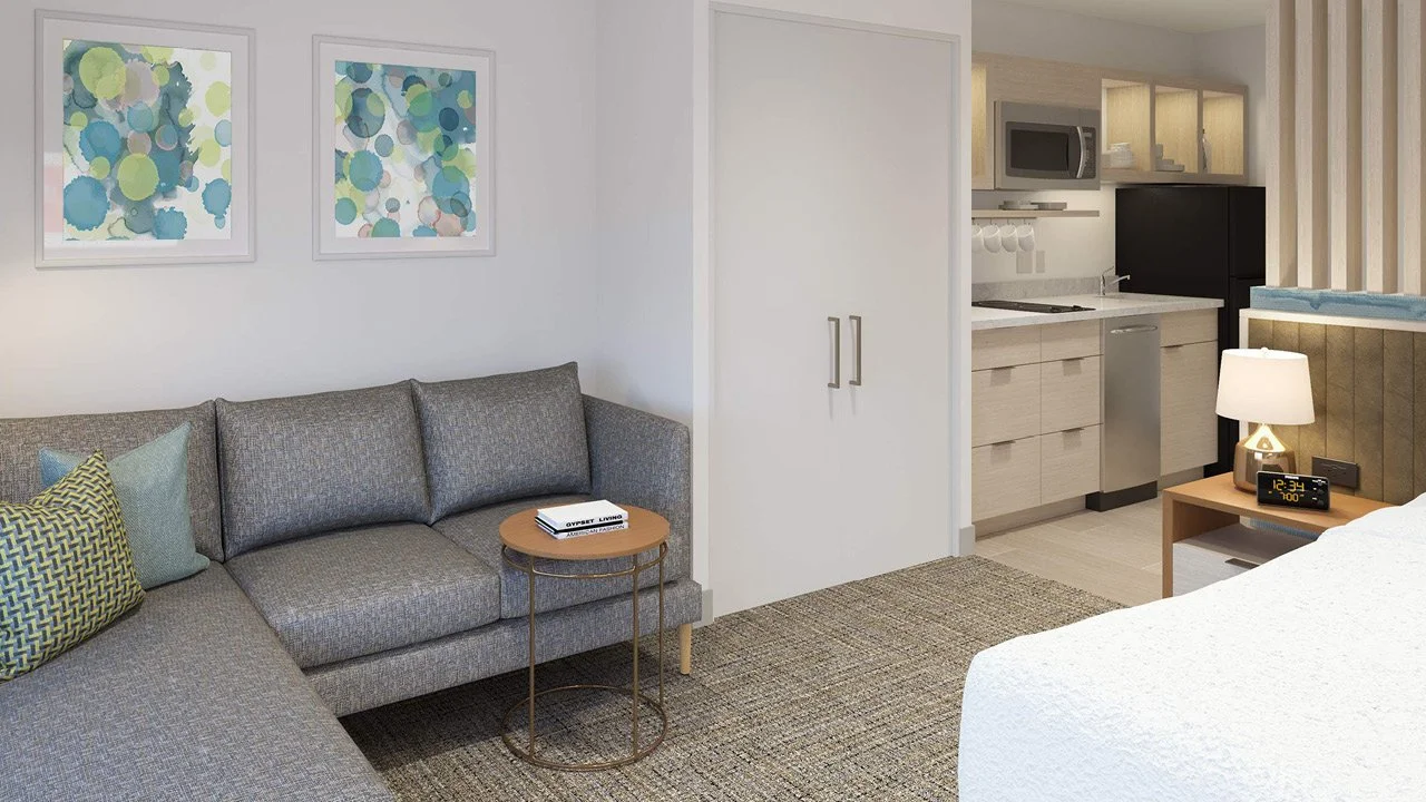 Towneplace Suites by Marriott hotel guest room with a grey sofa, colorful pillows, wall art, a small wooden side table, and a kitchen area with white cabinets, a microwave, and a bed with a white duvet and a nightstand with a lamp and clock.