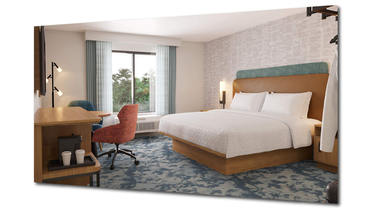 A prototypical hotel room for Hampton Inn by Hilton, with a large bed, a nightstand with a lamp, a window with curtains, and a sitting area with a desk and chairs.