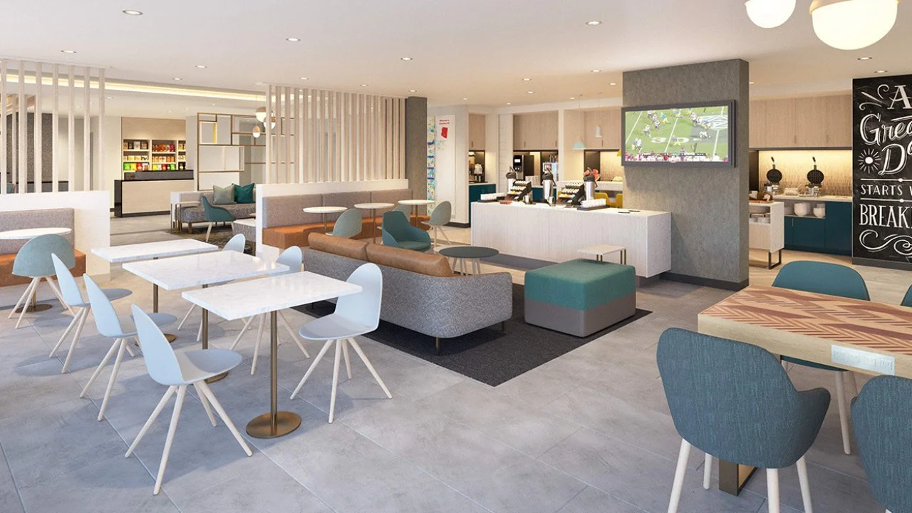 Towneplace Suites by Marriott prototypical hotel lobby with white tables, chairs with wooden legs, and various seating areas including sofas and armchairs. A TV is mounted on a pillar, and a chalkboard menu is visible in the background.