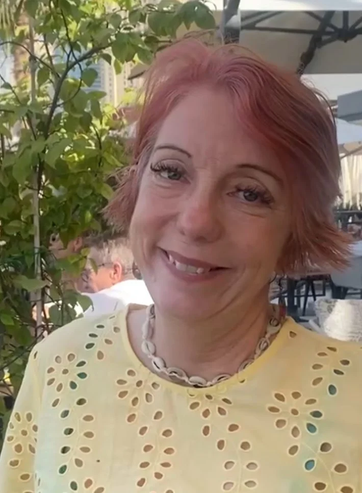 A woman with short red hair outdoors, smiling, wearing a yellow top with cut-out patterns and a shell necklace.