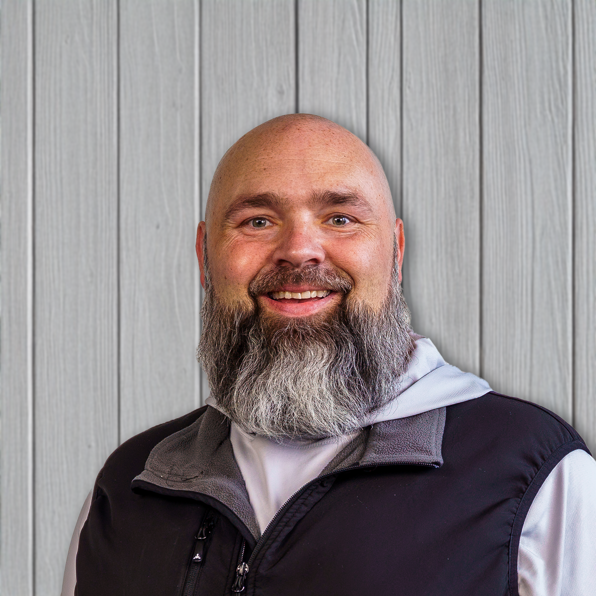 Portrait of a smiling, bald man with a full beard, wearing a black vest over a white shirt, standing in front of a light-colored wooden wall.