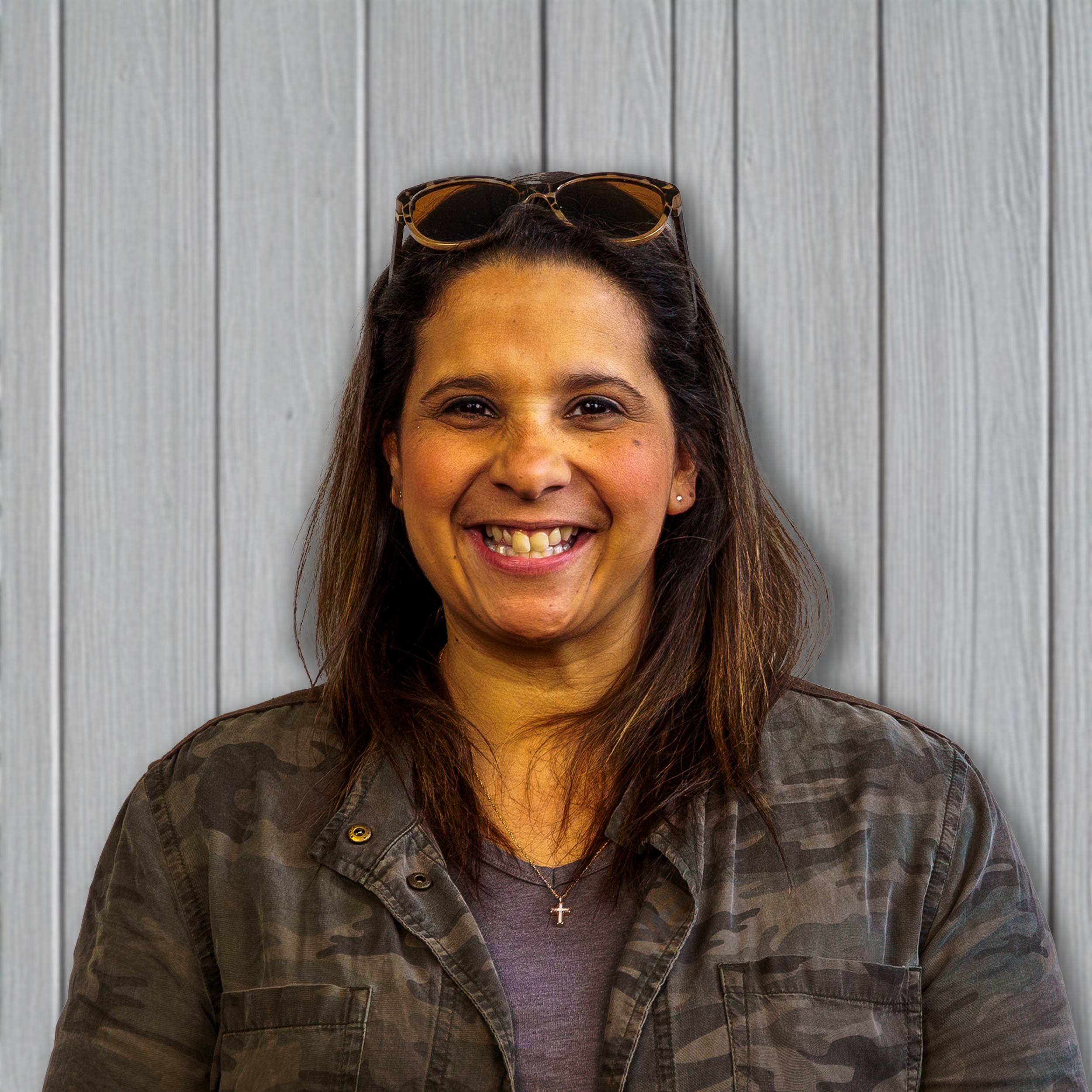 Smiling woman with glasses on head and camouflage jacket standing in front of a wooden wall