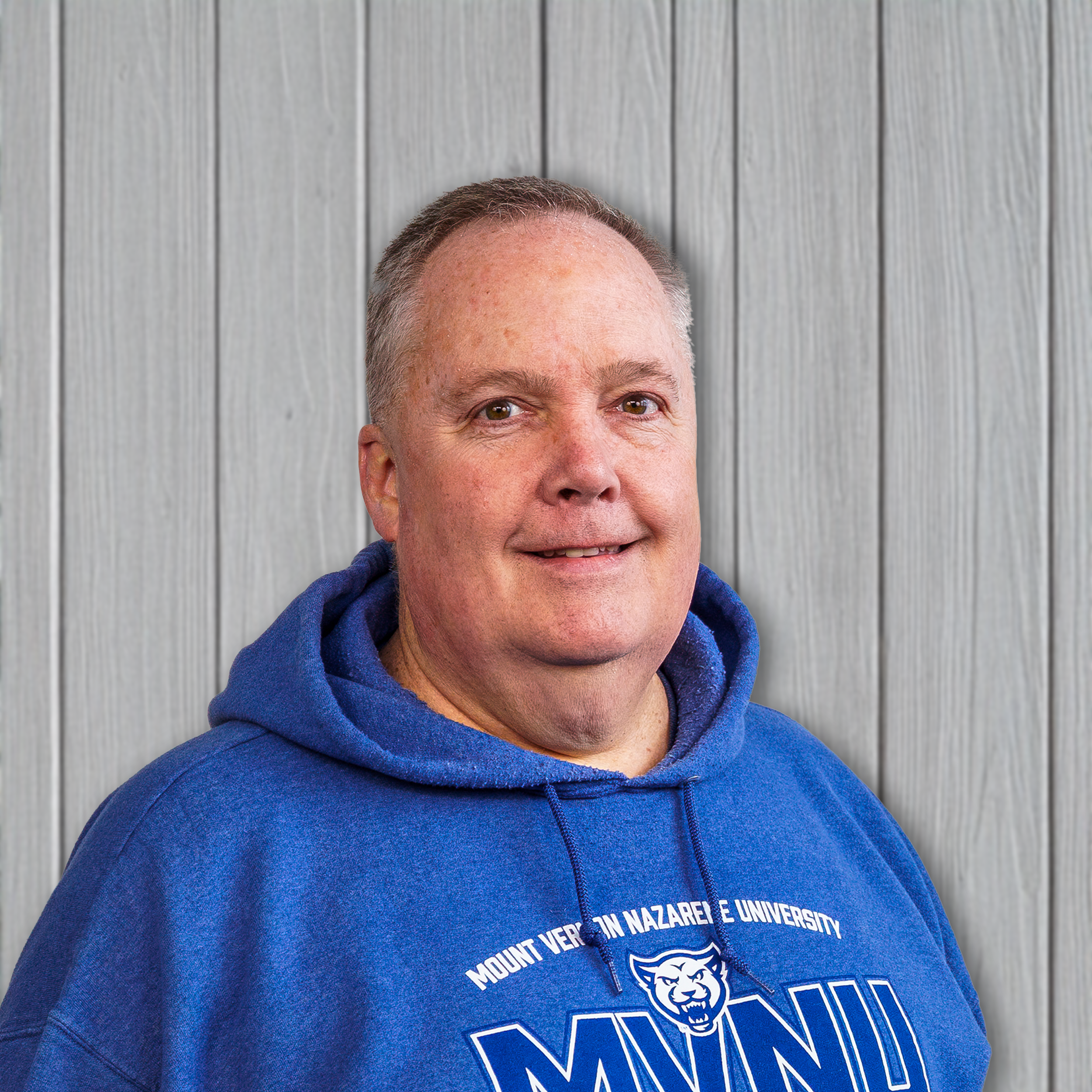 A middle-aged man with short gray hair, wearing a blue hoodie with Mount Vernon Nazarene University logo, standing against a light wood wall.