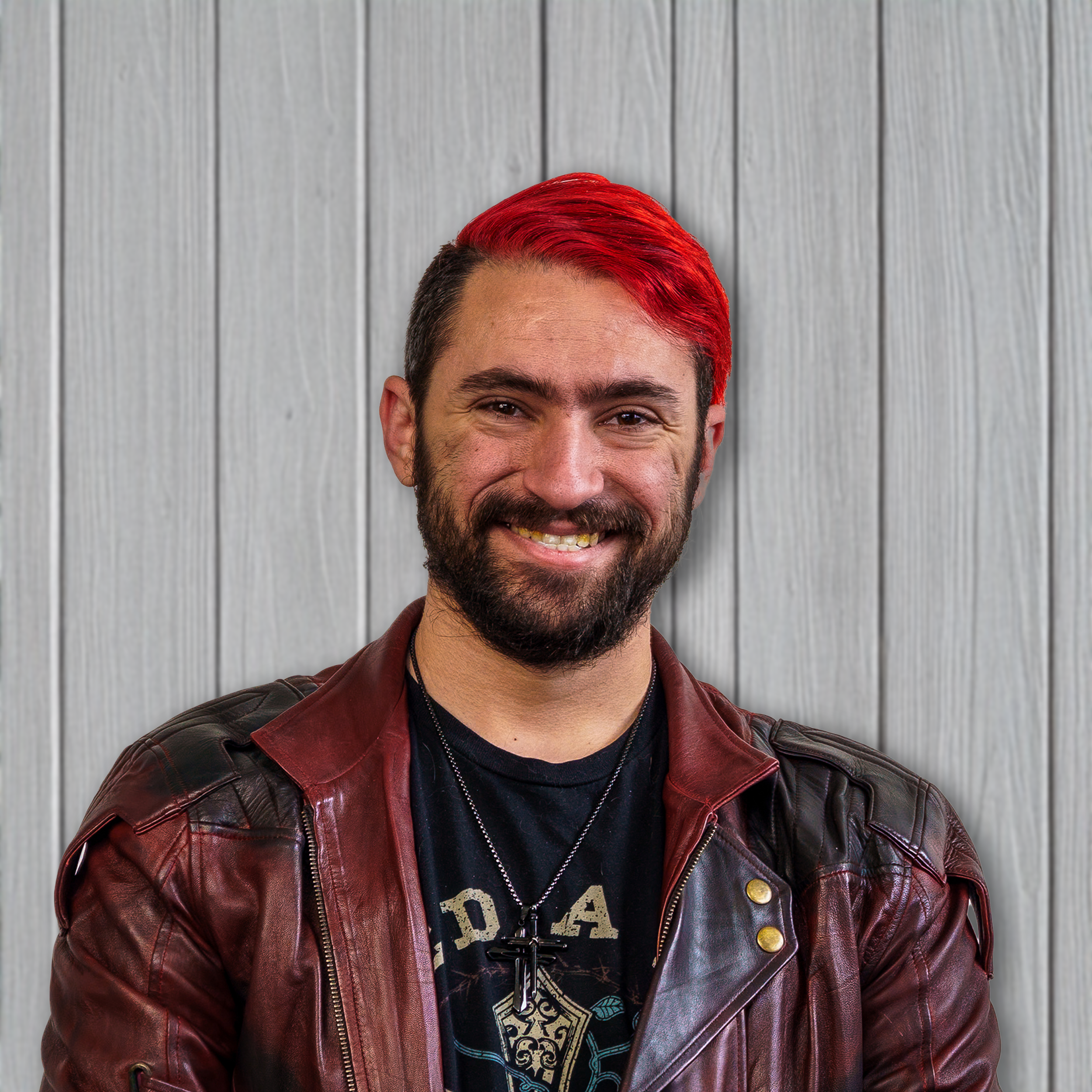 A man with a beard and red hair, smiling, wearing a black t-shirt and a red leather jacket, standing against a light gray wooden background.