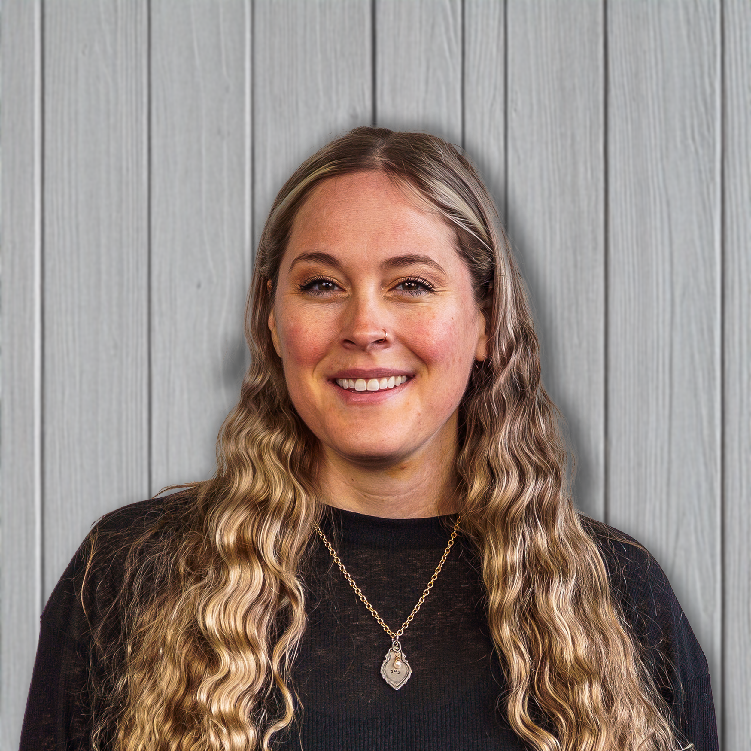 A young woman with long, wavy blonde hair smiling in front of a light wood-paneled background.