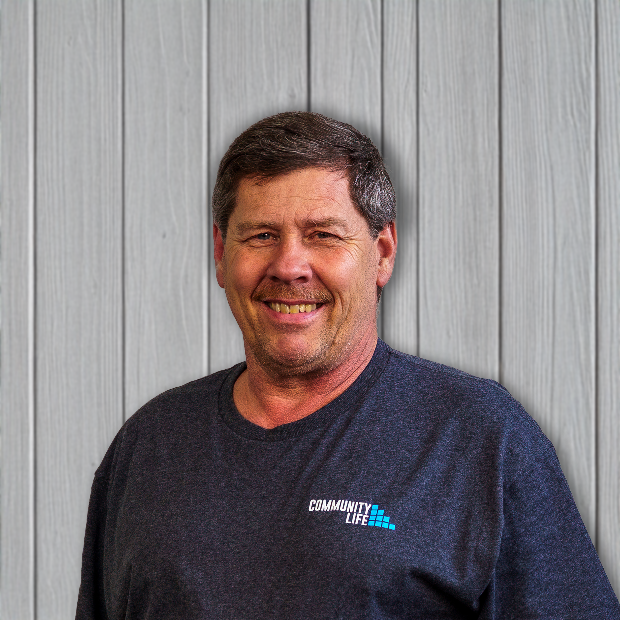 A man with short brown hair and a beard, smiling, standing in front of a wooden wall. He is wearing a dark gray t-shirt with a small logo that says 'Community Life' on the chest.