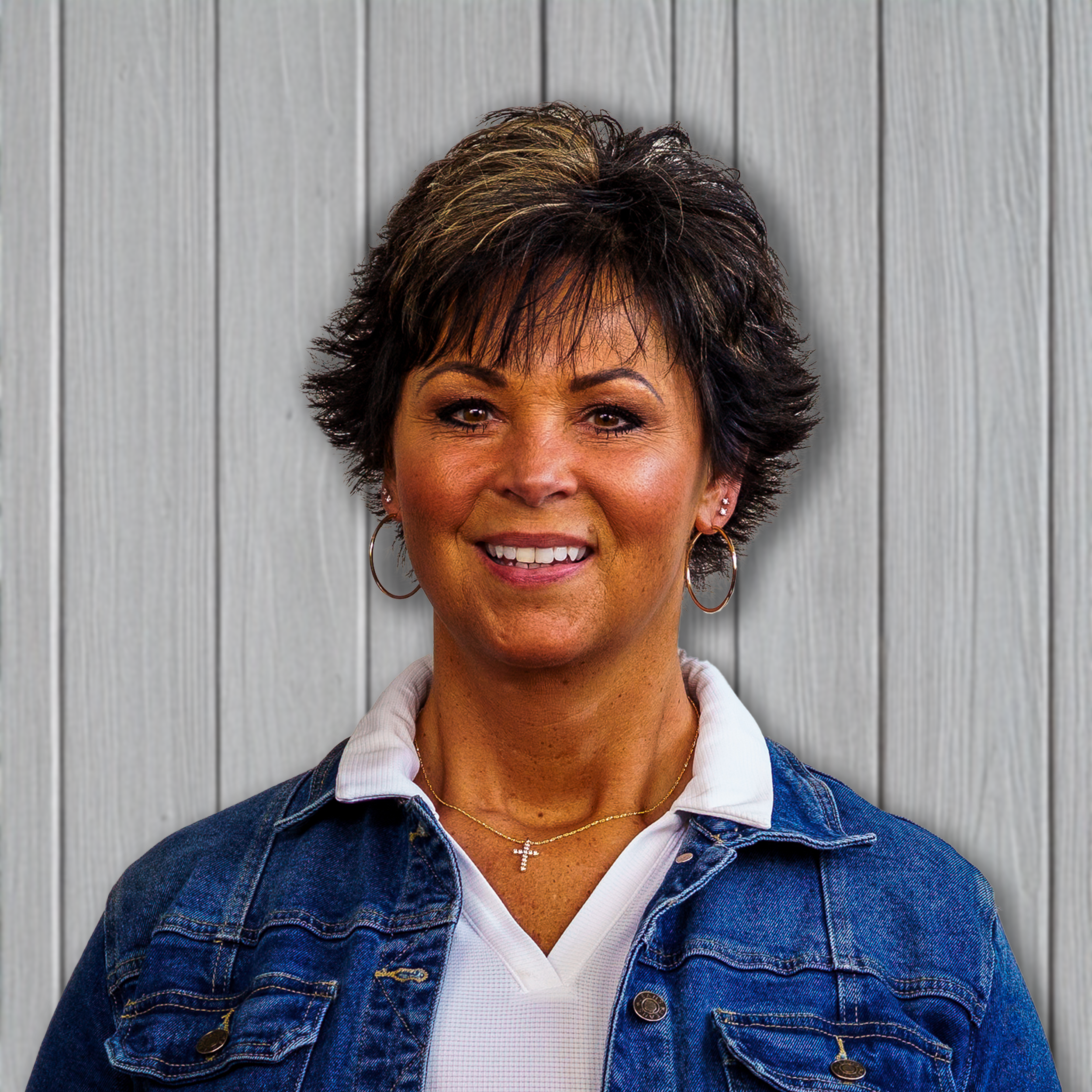 A woman with short dark hair and hoop earrings, wearing a white collared shirt and a denim jacket, standing in front of a light gray wooden wall.