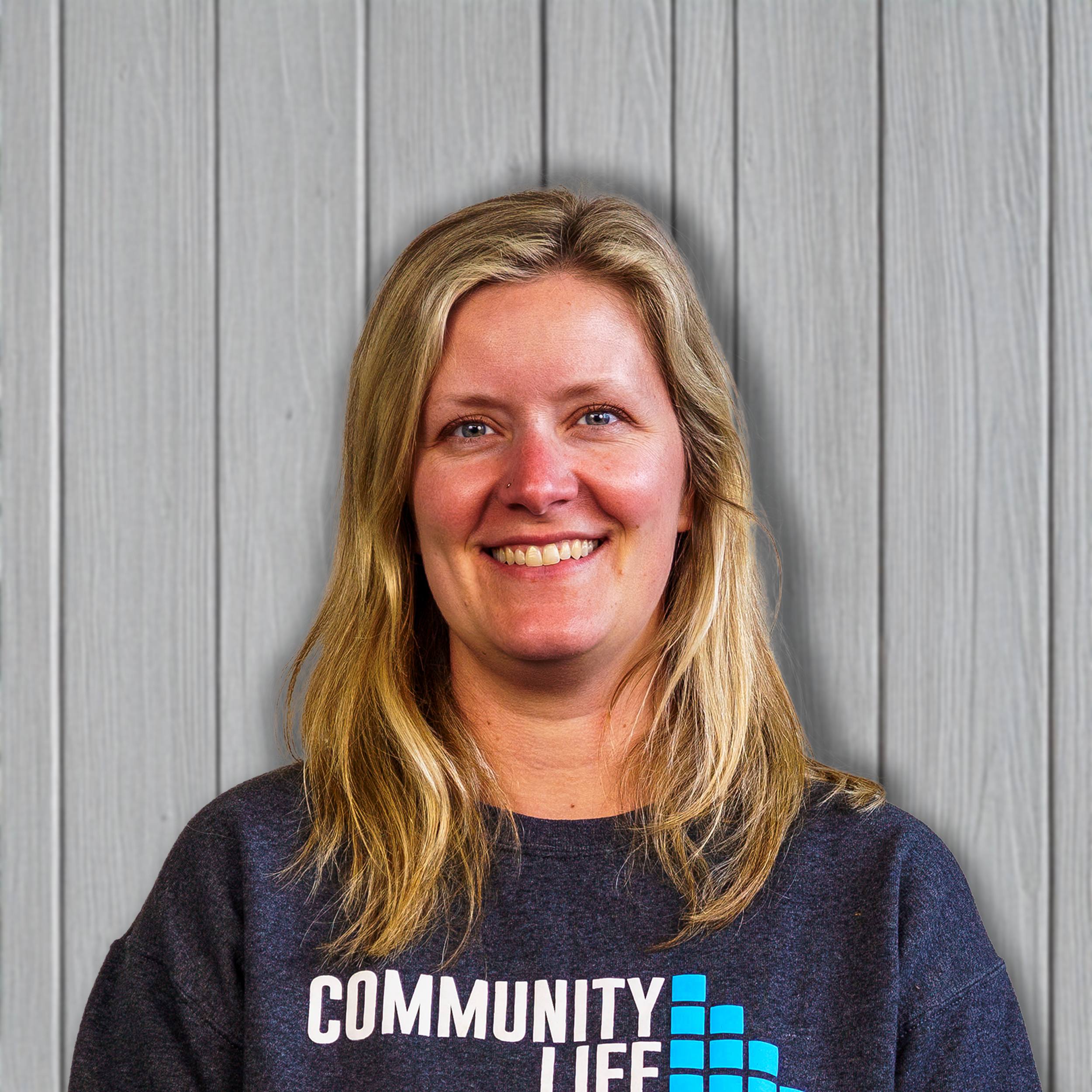 A smiling woman with blonde hair wearing a black T-shirt that says "Community Life" in white and blue text, standing in front of a light gray wooden panel background.