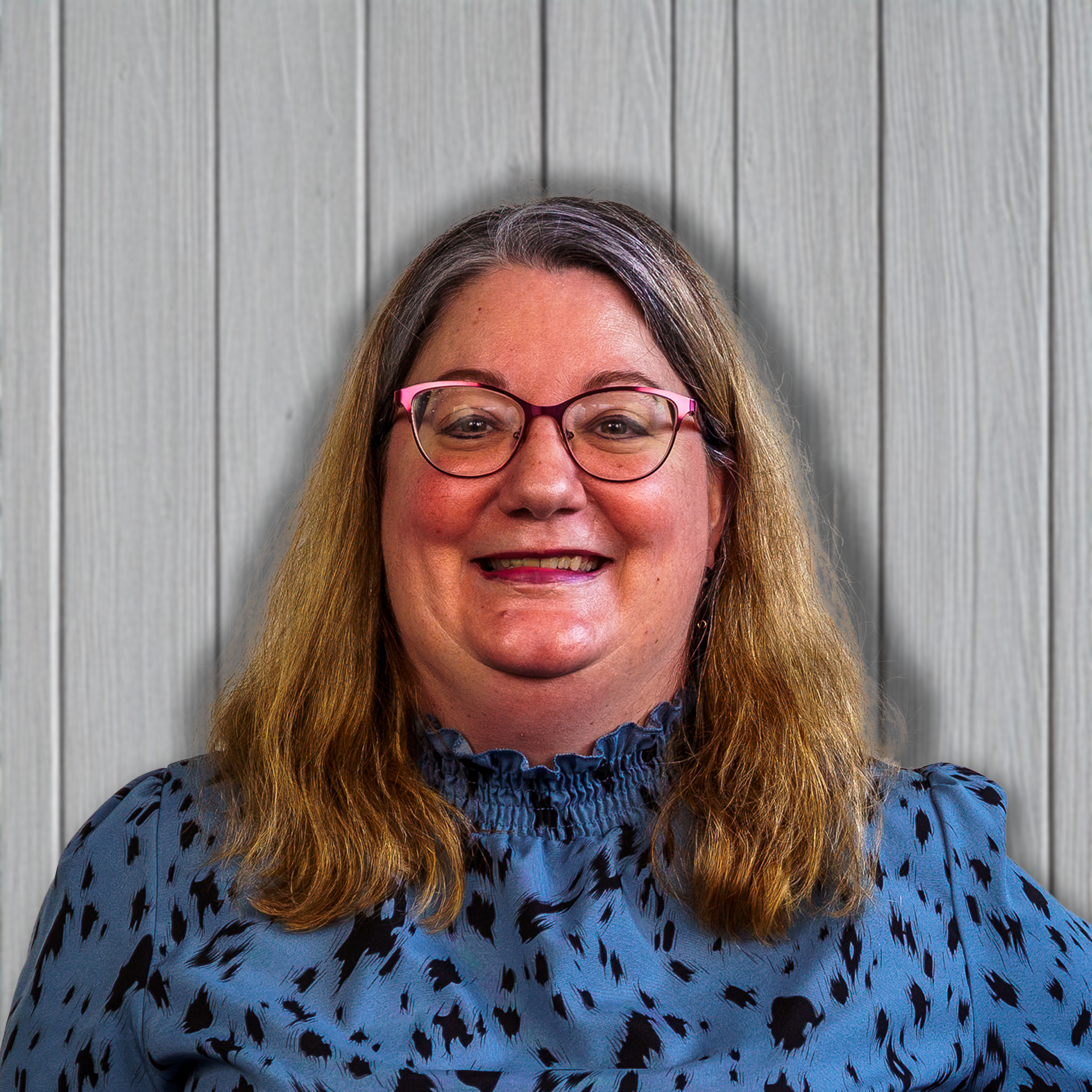 A woman with shoulder-length brown hair, wearing glasses with pink frames and a blue animal-print blouse, smiling in front of a light wooden wall.