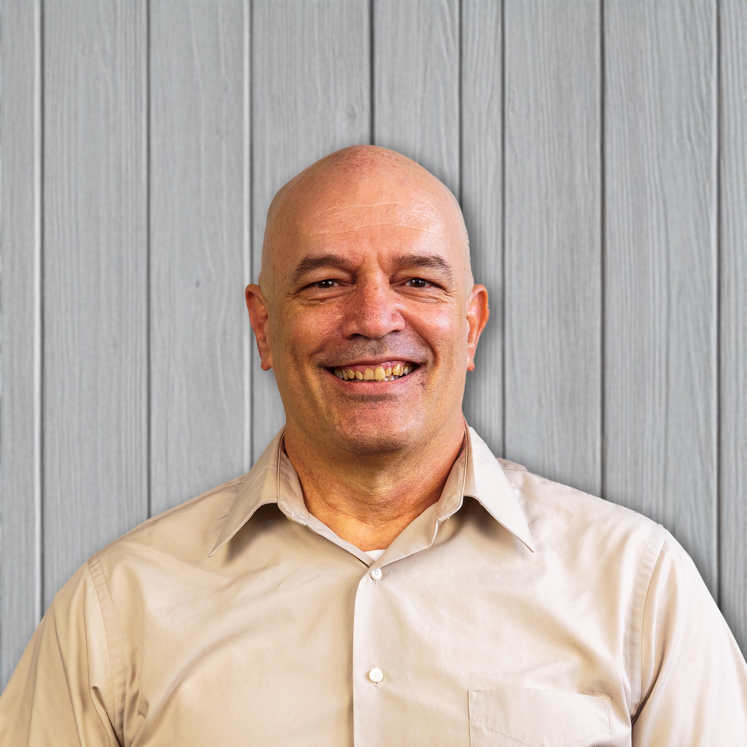 A bald man with a big smile wearing a beige button-up shirt, standing in front of a gray wooden wall.