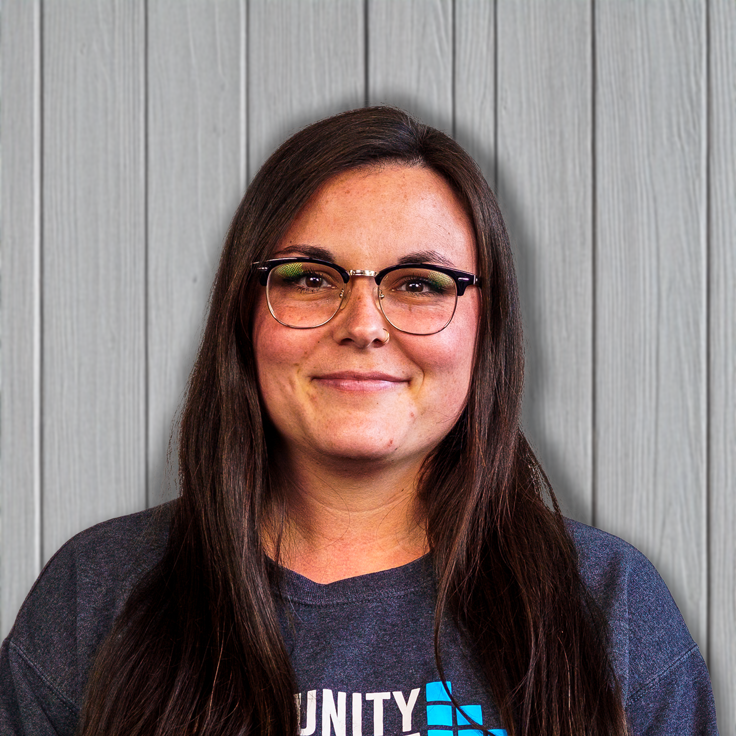 A woman with long brown hair, glasses, and a nose ring, smiling, standing against a light gray wooden wall.