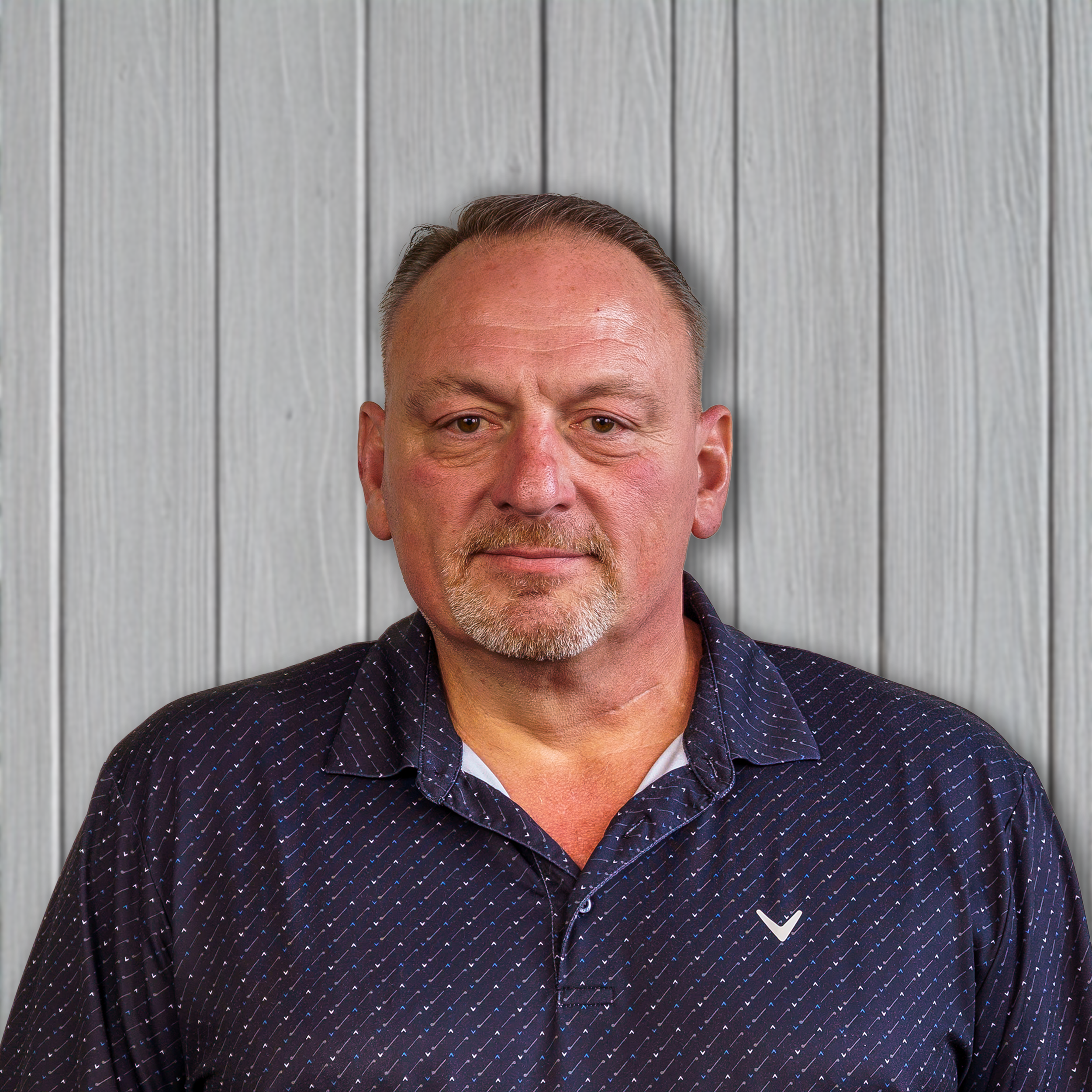 A middle-aged man with short hair, a goatee, wearing a dark patterned shirt, standing against a light gray wooden wall.