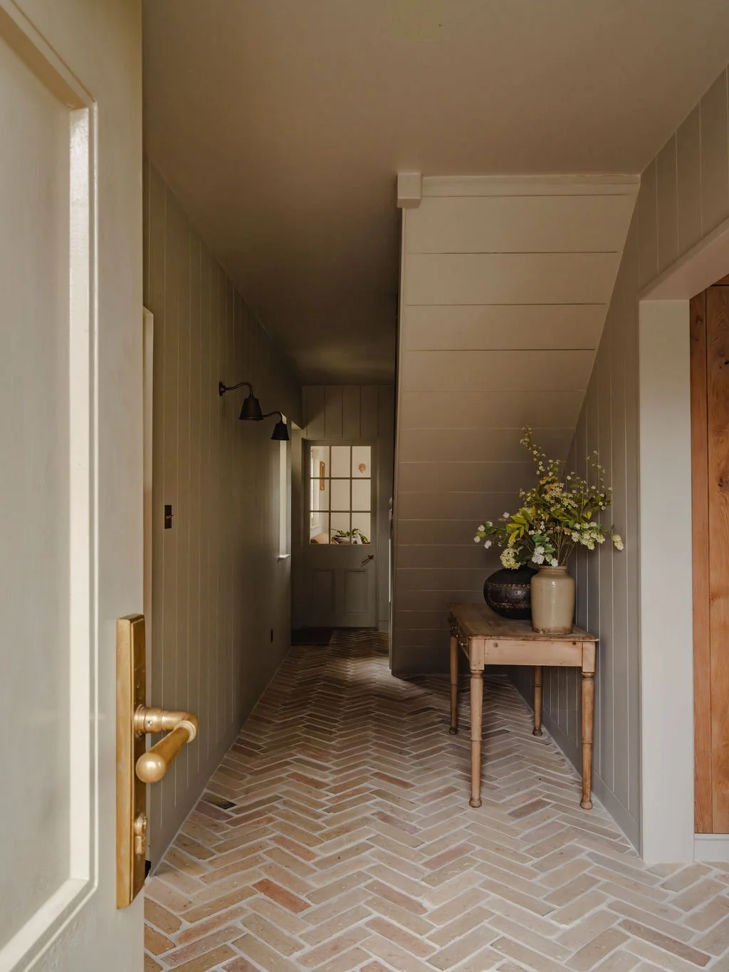 A country cottage hallway with a rustic reclaimed herringbone brick floor, beige panneled walls, a small wooden table with two vases and flowers, and a door at the end of the hallway.