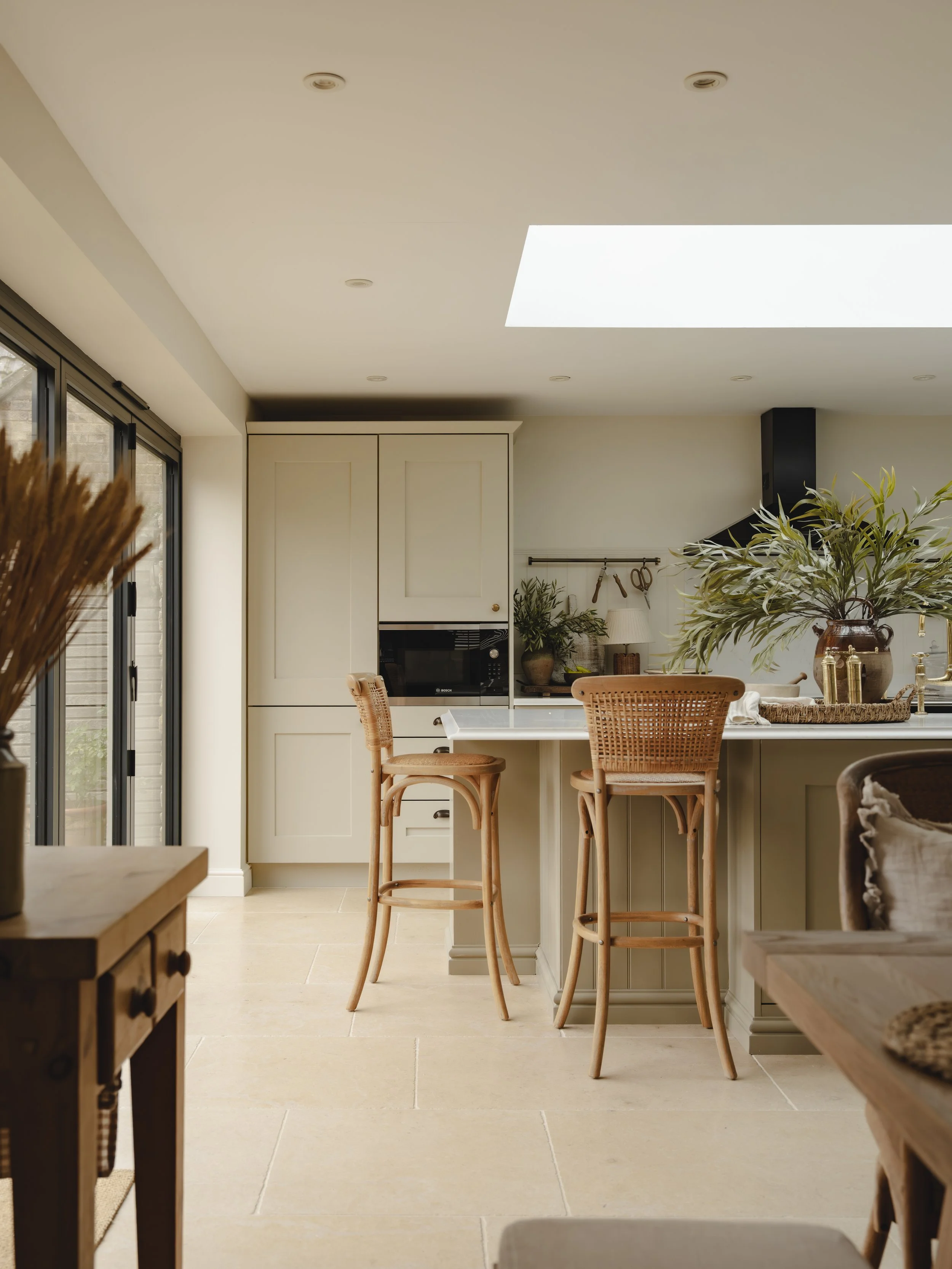 Country kitchen with limestone flagstone floor. Bright kitchen with beige cabinetry, a white quartz island with rustic bar stools, large potted plants, and a skylight letting in natural light.