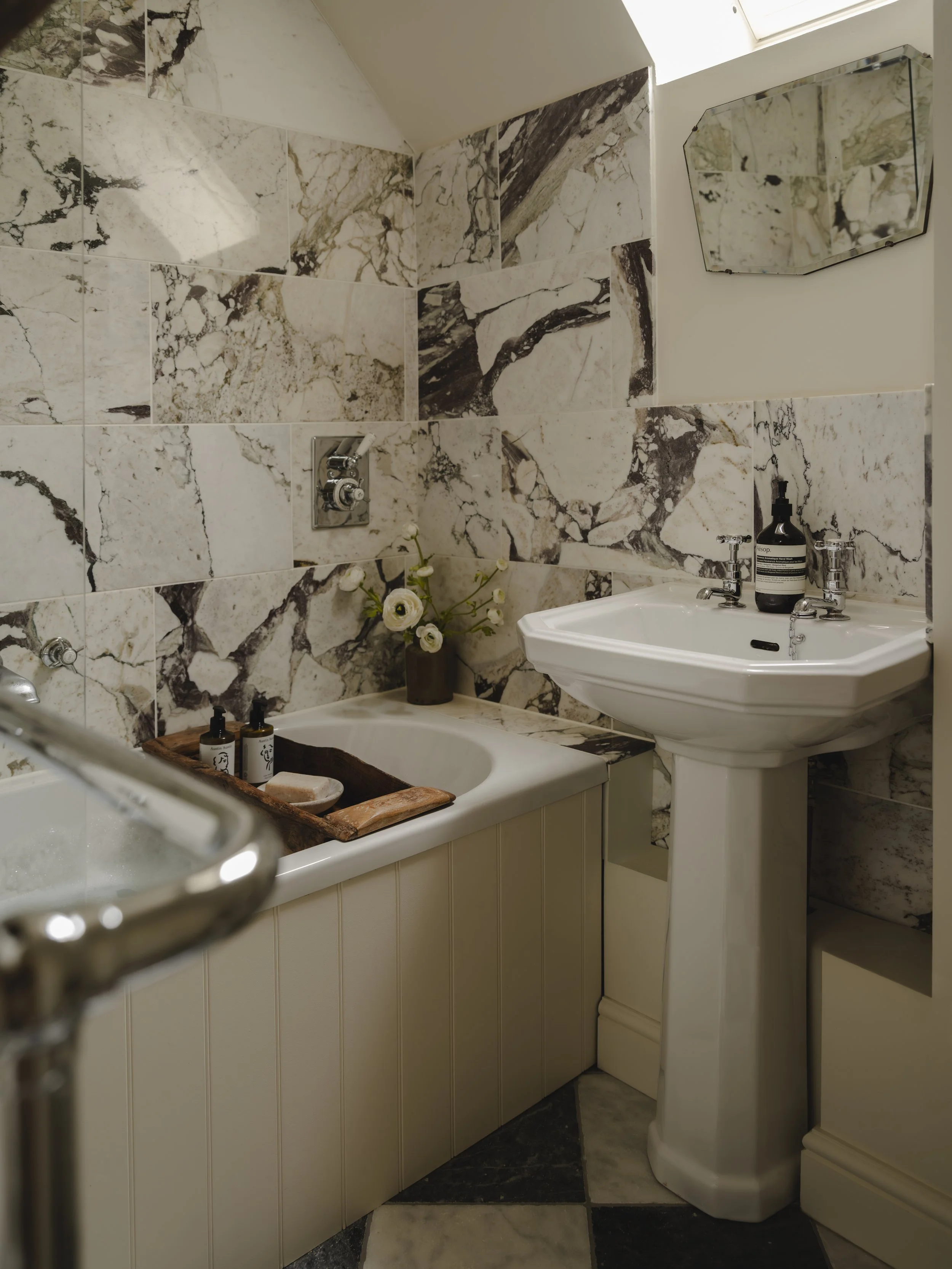A small bathroom with a marble tiled wall, a pedestal sink with vintage faucet fixtures, a mirror above the sink, a bathtub with a wooden tray holding bottles, a vase with white flowers, and a window providing natural light.