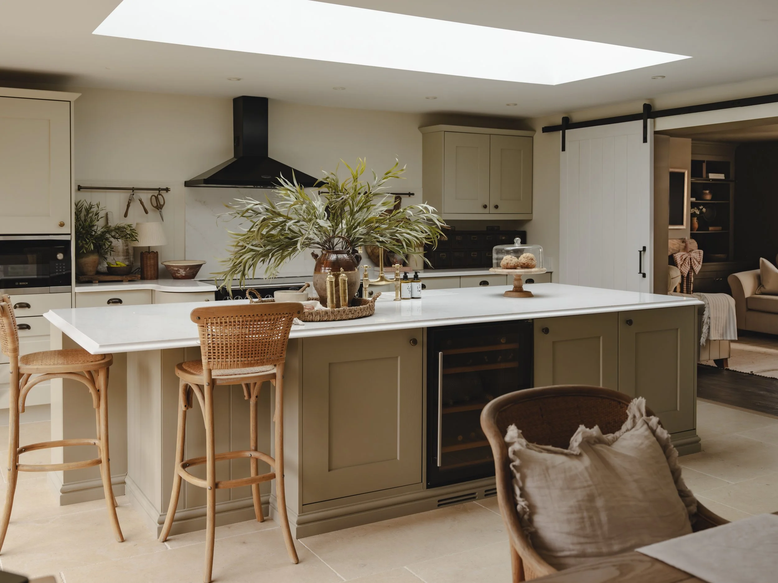 Modern kitchen with beige cabinets, a large white central island with a wine cooler, wicker bar stools, and a large plant in a brown pot as decoration.
