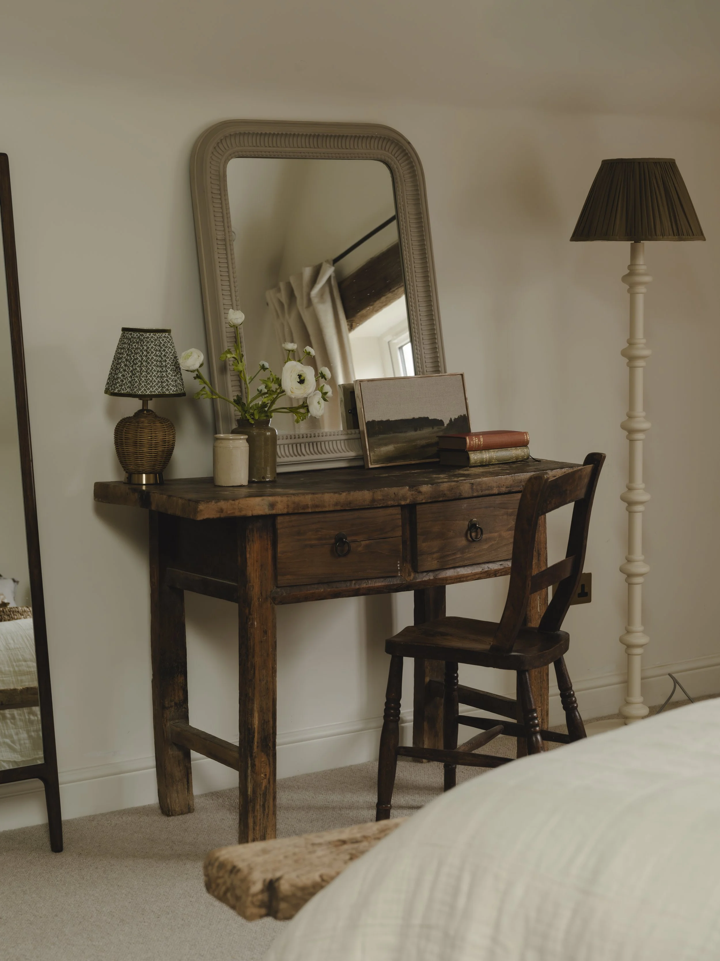 Rustic Wooden desk dressing table with a mirror, a vase of white flowers, and some books, positioned next to a cream-colored bed with a bench at the foot, in a cozy bedroom.