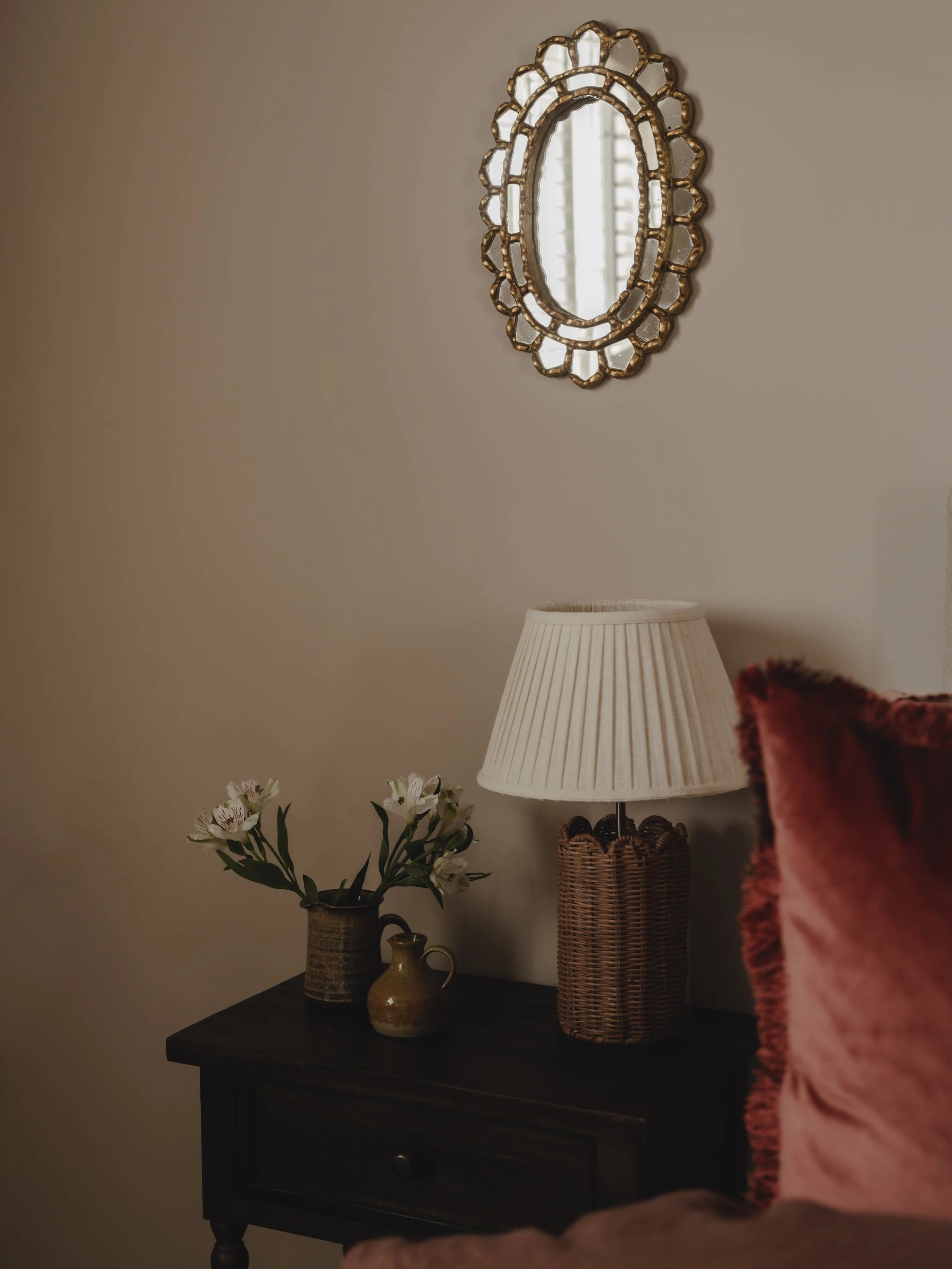 A bedroom corner with a dark wood nightstand holding a vase of white flowers, a small ceramic pitcher, and a woven lamp with a beige lamp shade. Above, a decorative antique oval mirror with a gold ornate frame is on the beige wall.