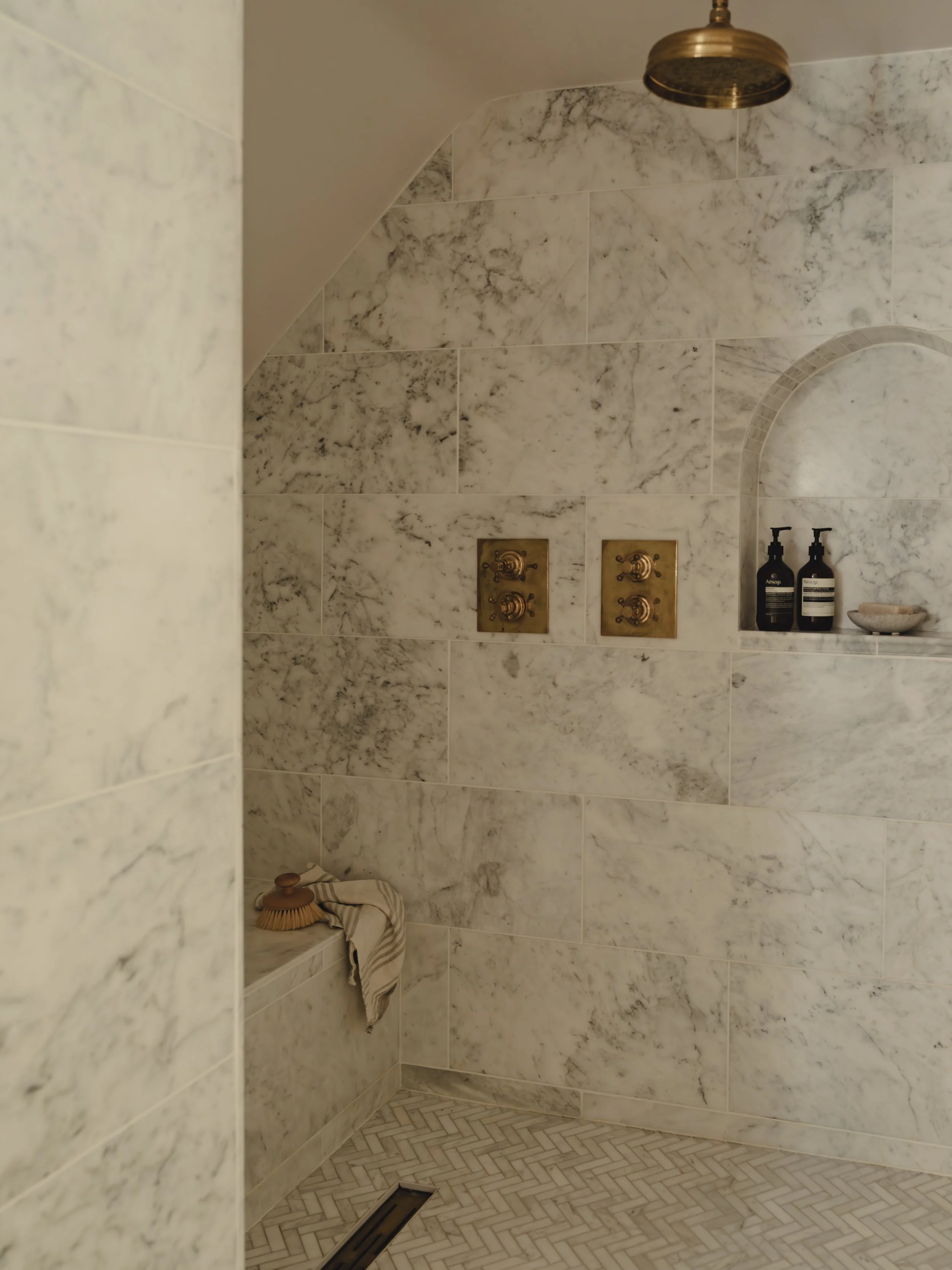 A luxurious marble-tiled shower area with arched shower niche, a brass showerhead, and brass control valves. A wooden scrub brush and towel are on a ledge in the corner. The shower floor has a herringbone marble tile pattern.