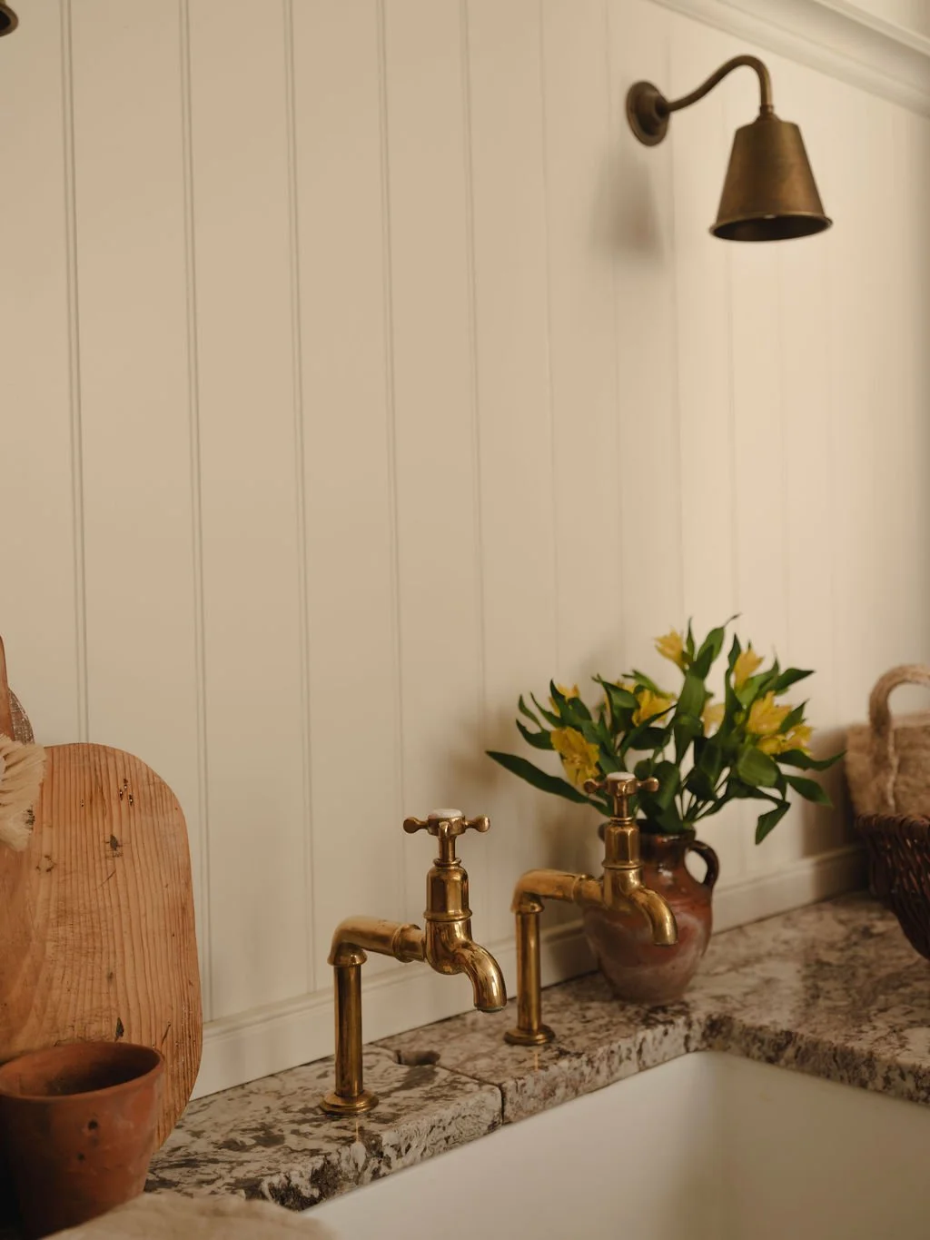 Kitchen sink with brass faucets, a vase of yellow flowers, a wooden cutting board, and a small clay pot on a granite countertop, with a wall-mounted brass sconce light above.