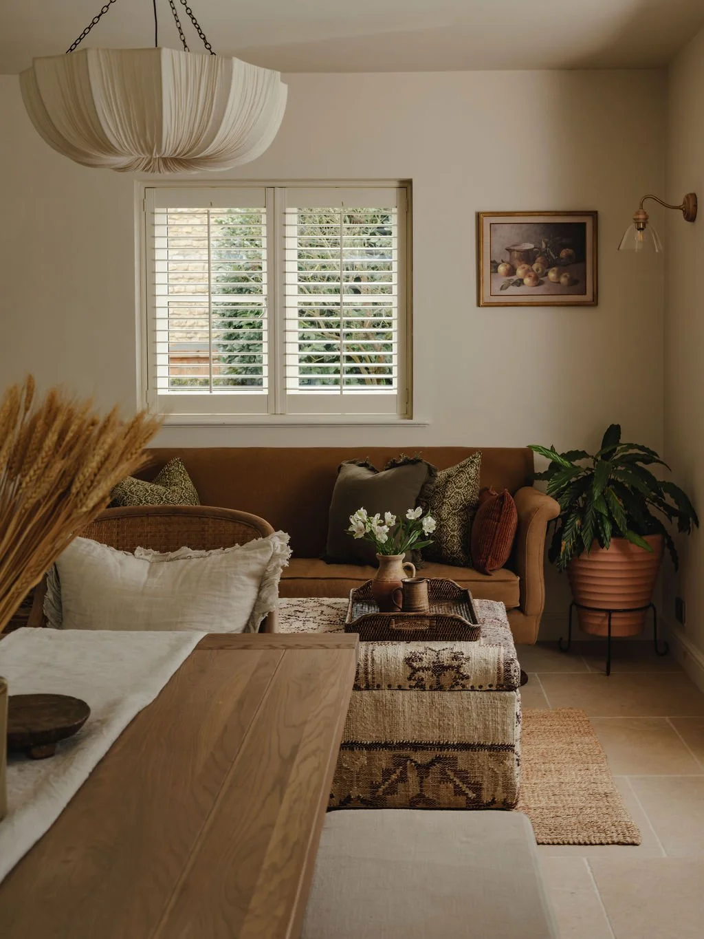 A cozy living room with a brown sofa, decorative pillows, a wooden coffee table, and a potted plant. There's a large window with white shutters, a framed Still Life painting on the wall, and a hanging ceiling light fixture.
