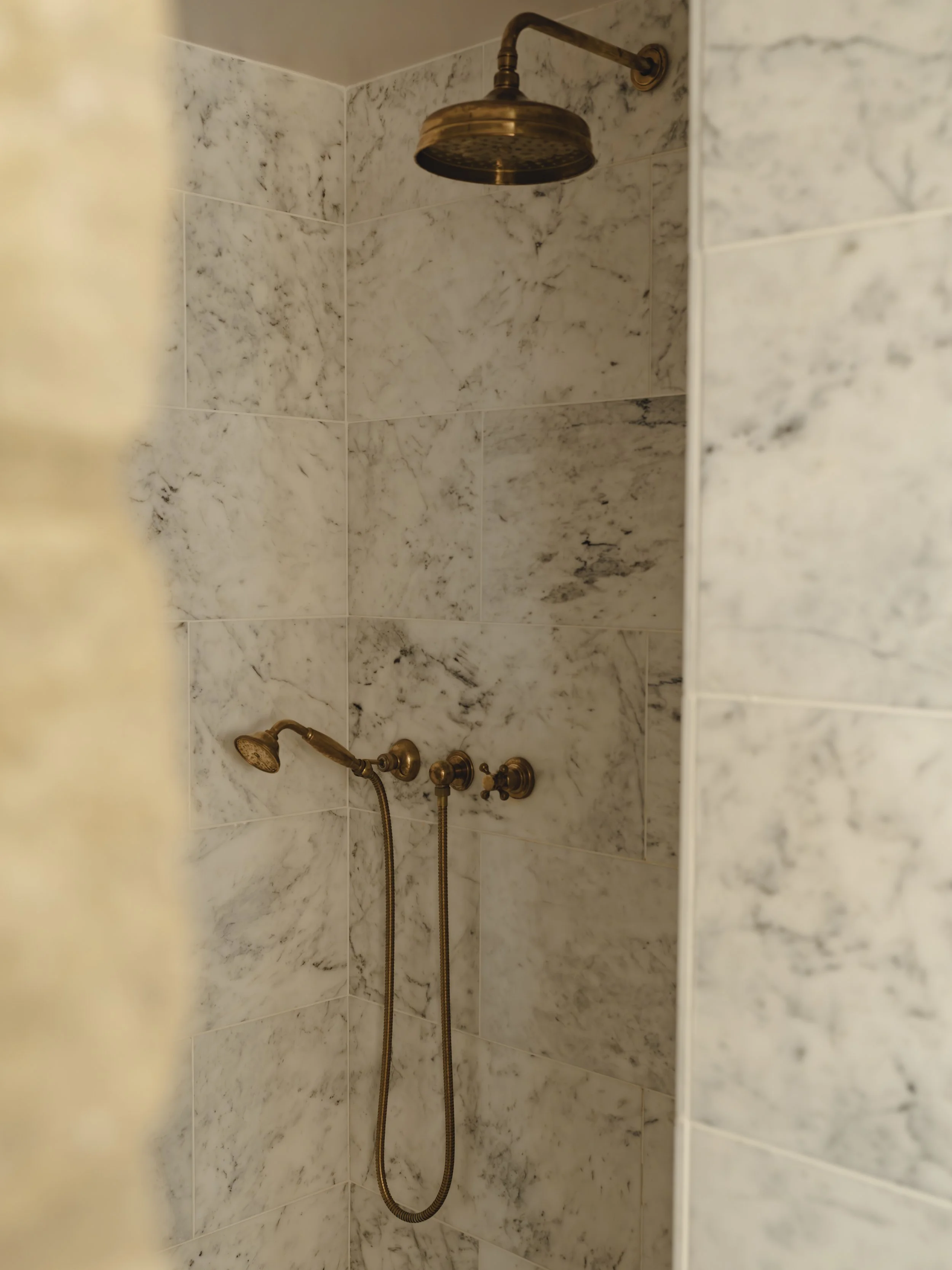 Marble shower with gold brass fixtures, including a rain showerhead and handheld shower, in a bathroom.