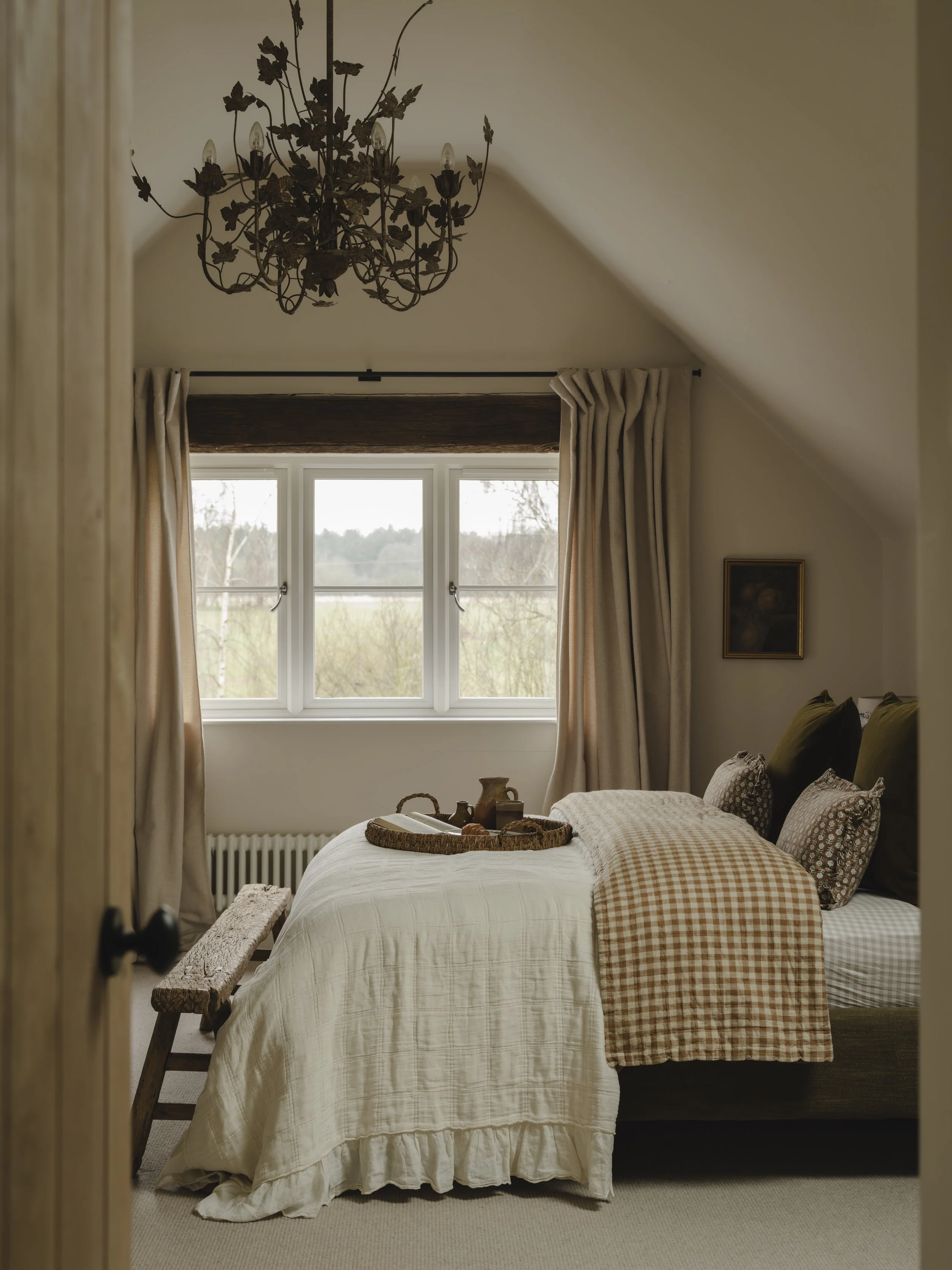 Cozy bedroom with a countryside view of trees and fields, cream-colored curtains, a rustic acorn leaf chandelier, and a bed with earthy-toned pillows and blankets, with a tray holding a teapot and cups.