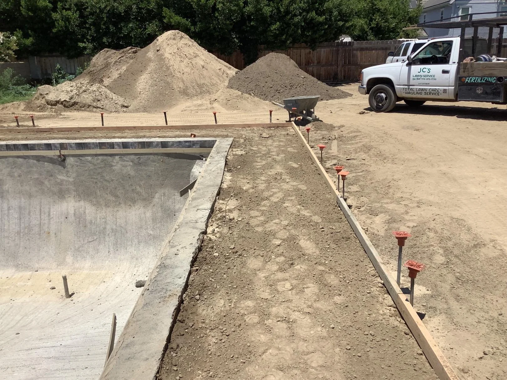 Construction site with a pool being built, dirt and sand piles in the background, a pickup truck parked nearby, and orange construction anchors along the edge.