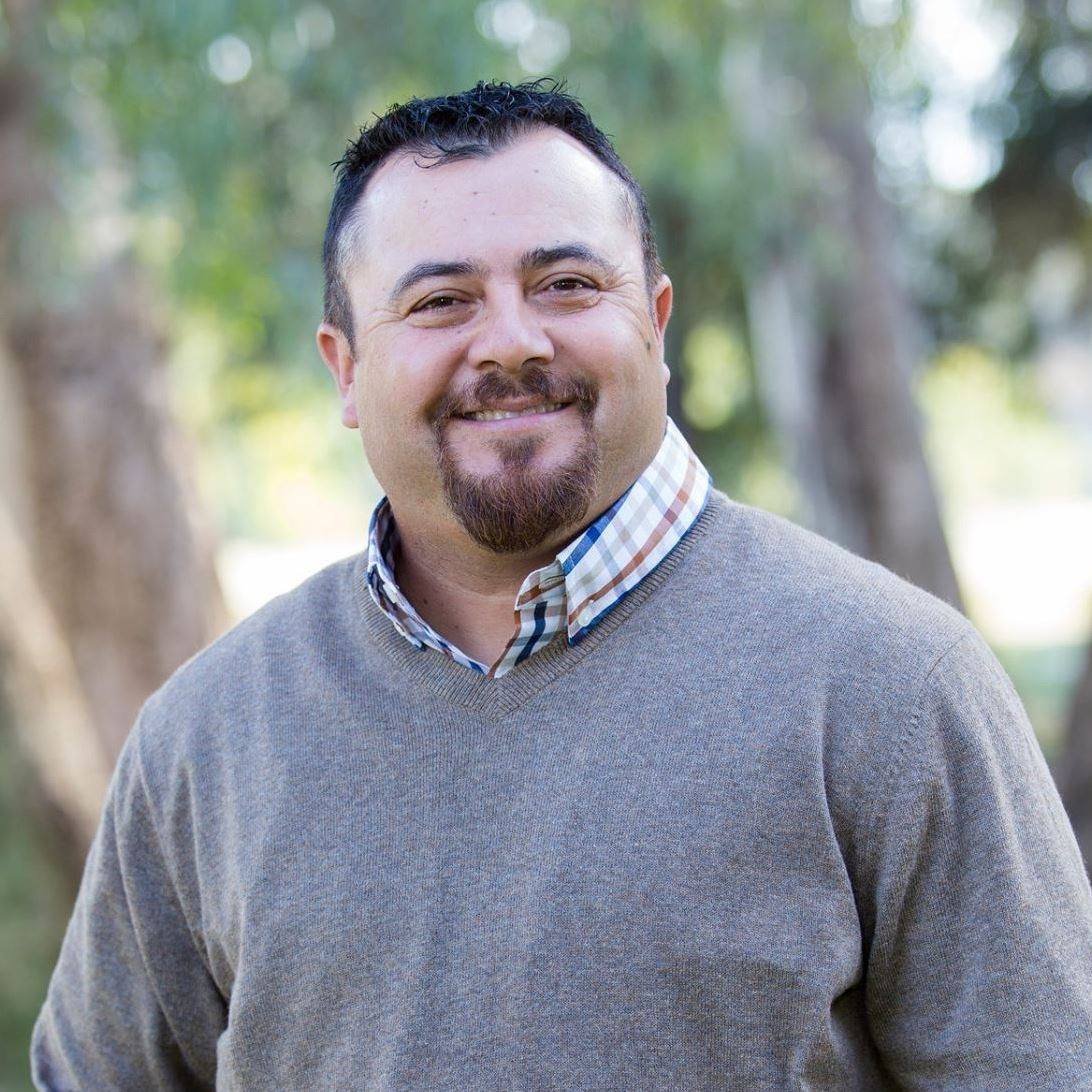 A smiling man with short dark hair and a goatee, wearing a gray sweater over a plaid collared shirt, outdoors with trees in the background.
